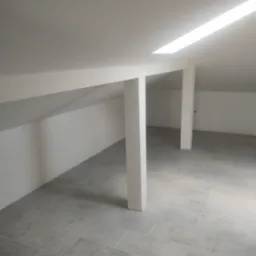 Poddasze po remoncie, widoczne białe ściany ze skosami, szare płytki podłogowe, białe filary podtrzymujące dach i wbudowane oświetlenie sufitowe.