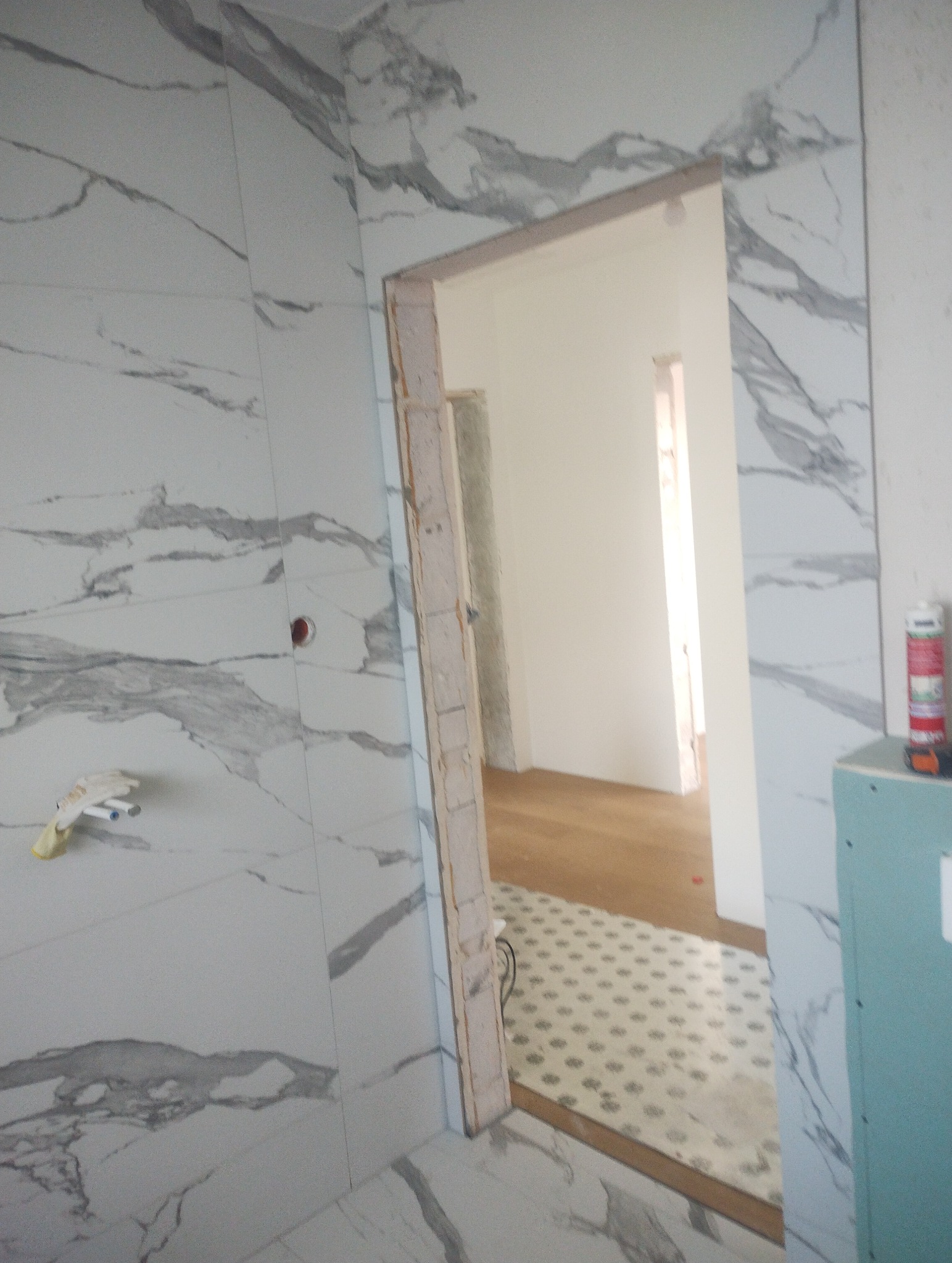 Wnętrze łazienki w trakcie remontu: ściany wyłożone płytkami imitującymi marmur, widoczna surowa ościeżnica drzwi, w tle podłoga z panelami i płytkami. Częściowo zamontowana płyta gipsowo-kartonowa.