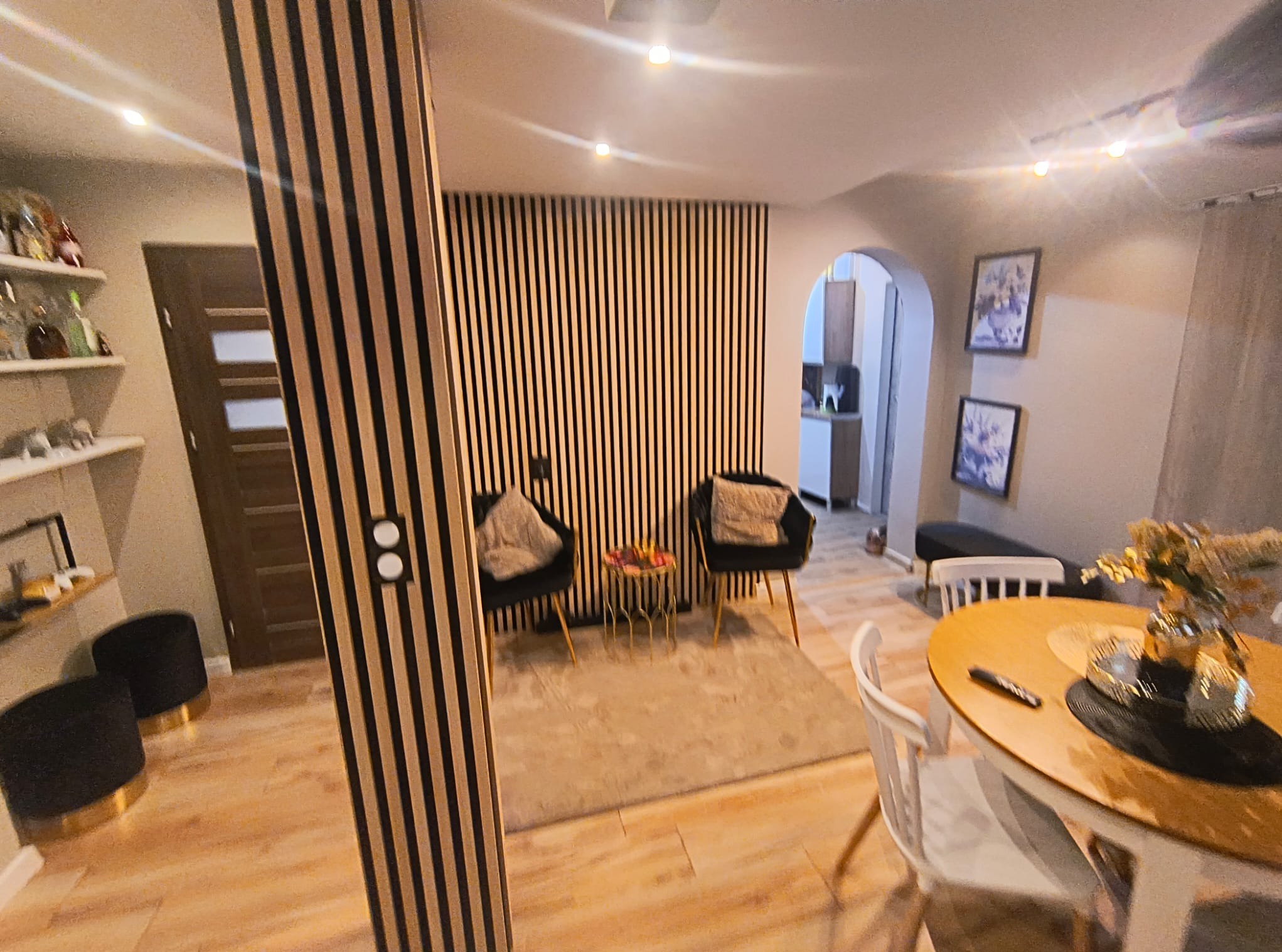 Salon z drewnianymi lamelami na ścianie, dwiema czarnymi fotelami, okrągłym stolikiem kawowym i dywanem w neutralnych barwach. Na pierwszym planie okrągły stół z białymi krzesłami i dekoracją.