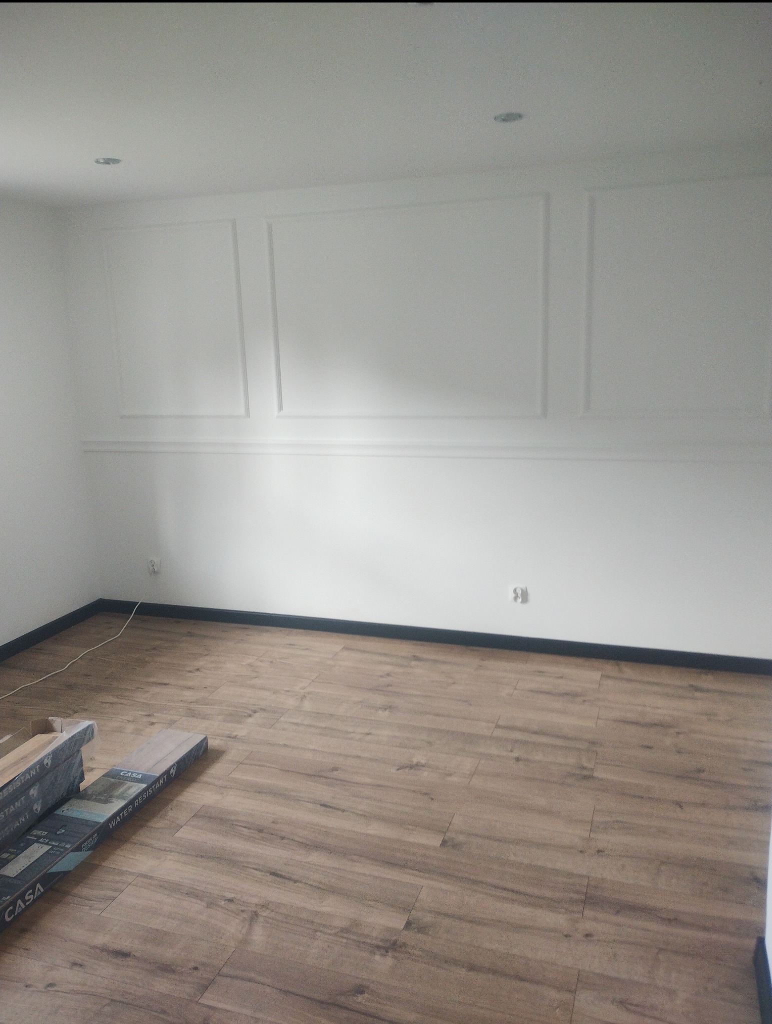 Pokój po remoncie z nową podłogą z paneli, widoczne paczki paneli pod ścianą, ściany pomalowane na biało z dekoracyjnymi listwami.