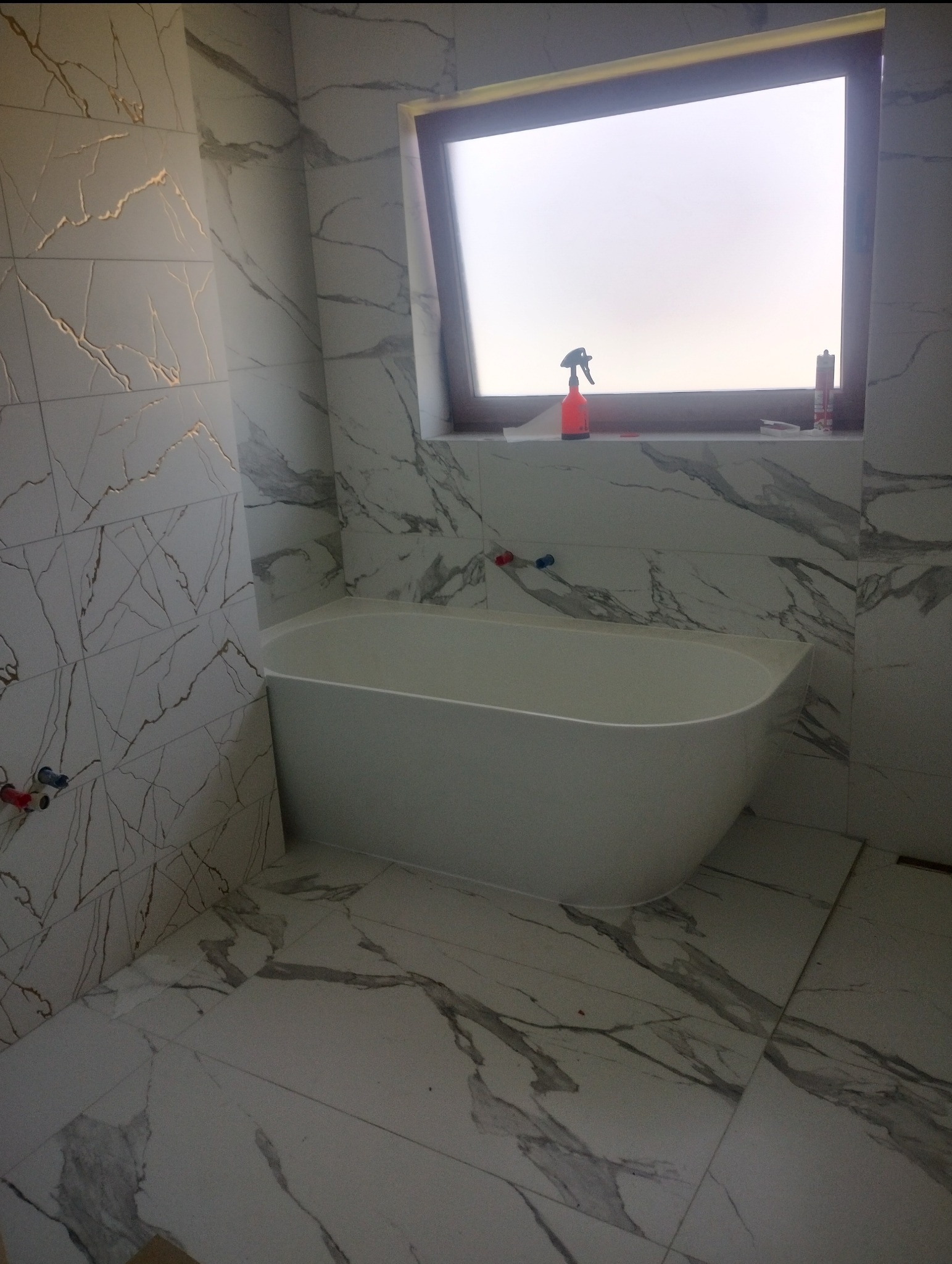 Nowoczesna biała wanna wolnostojąca w łazience wyłożonej płytkami imitującymi marmur, widoczne przyłącza wodne i akcesoria na parapecie okna.