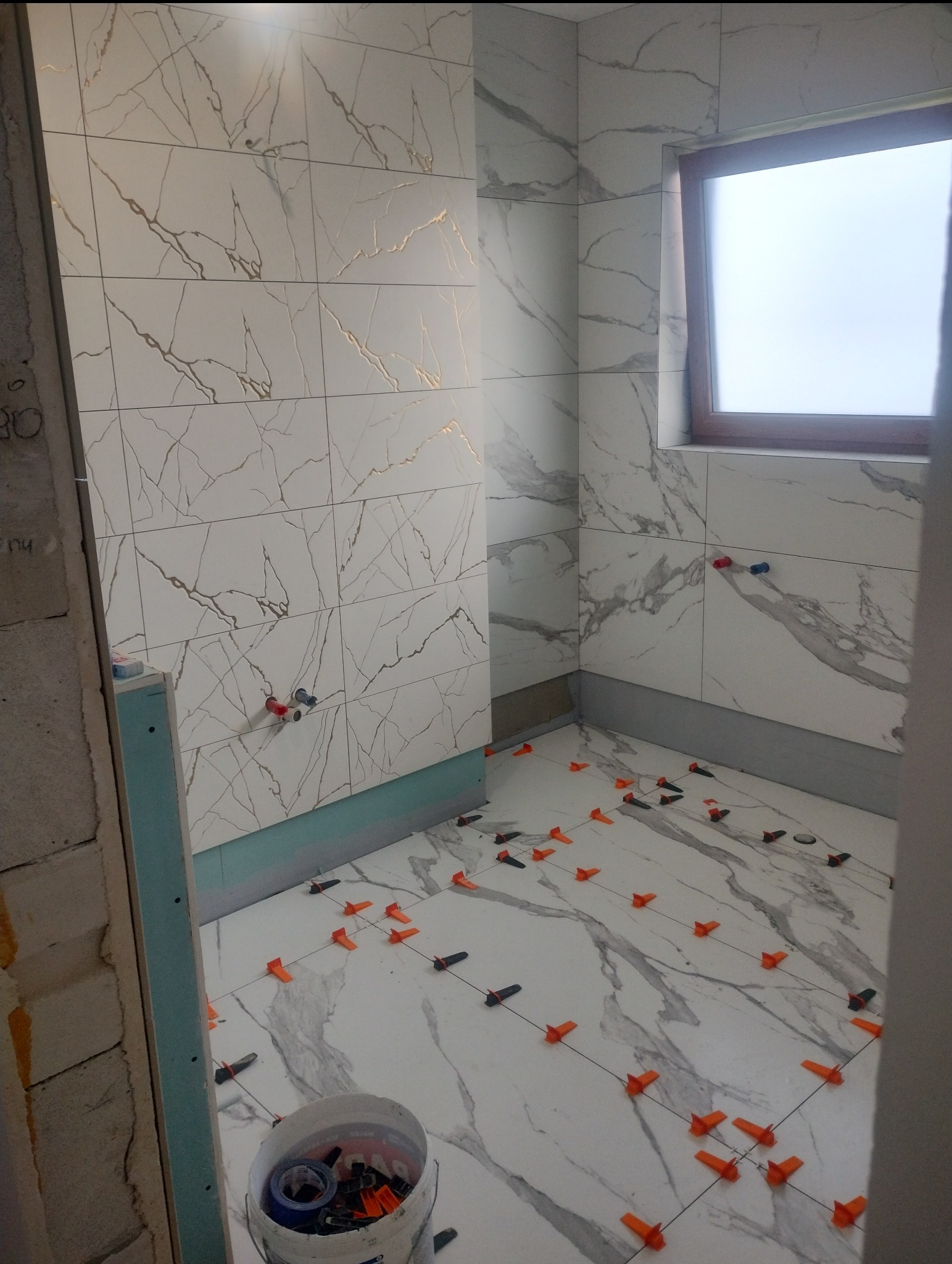 Łazienka w trakcie remontu w Koszalinie: białe płytki imitujące marmur na ścianach i podłodze, system poziomowania płytek, widoczne przyłącza wodne, kubeł z akcesoriami.