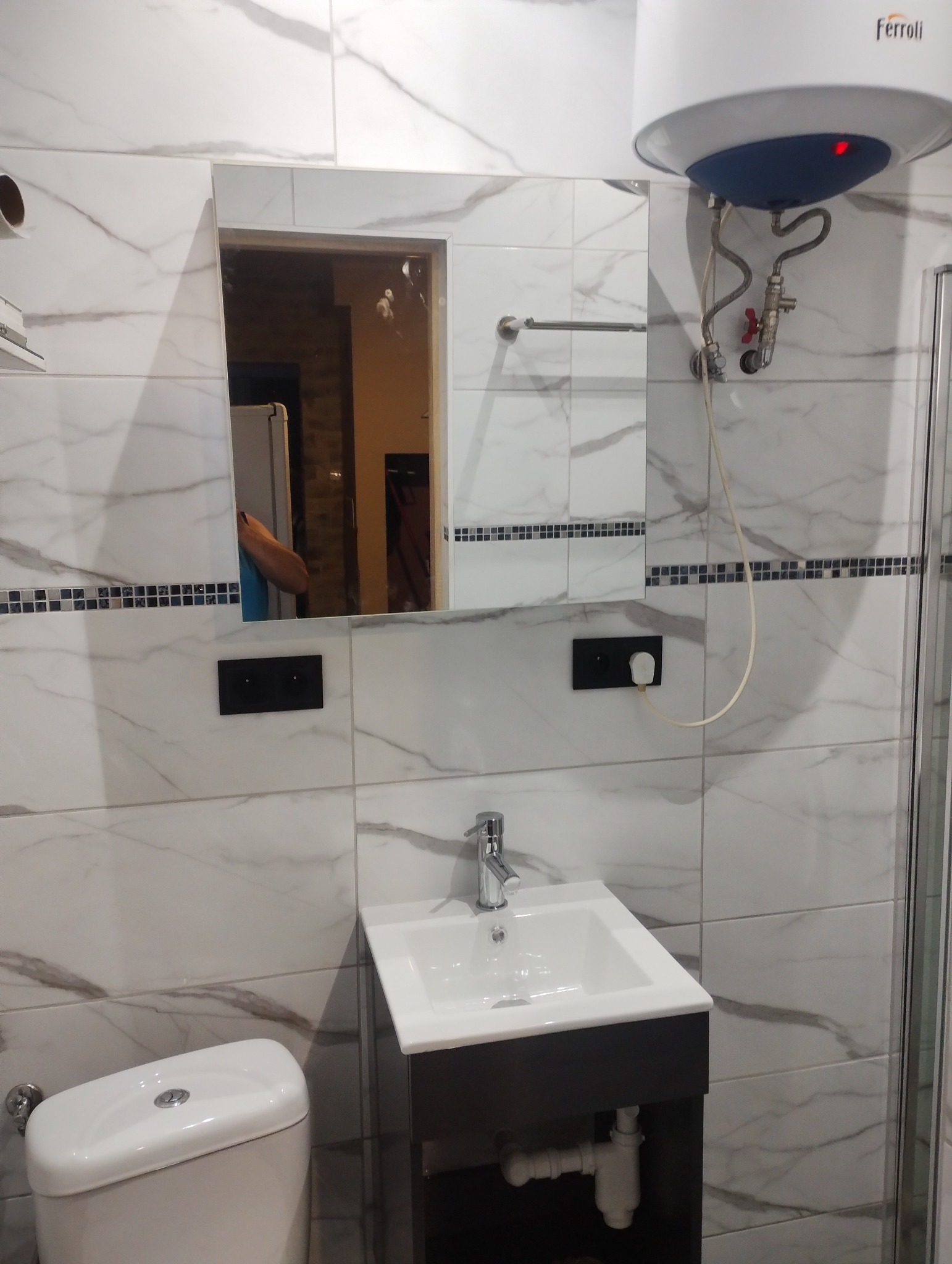 Nowoczesna łazienka z białymi płytkami imitującymi marmur, wisząca szafka z umywalką, toaleta i bojler elektryczny Ferroli. W lustrze odbija się fragment innego pomieszczenia.