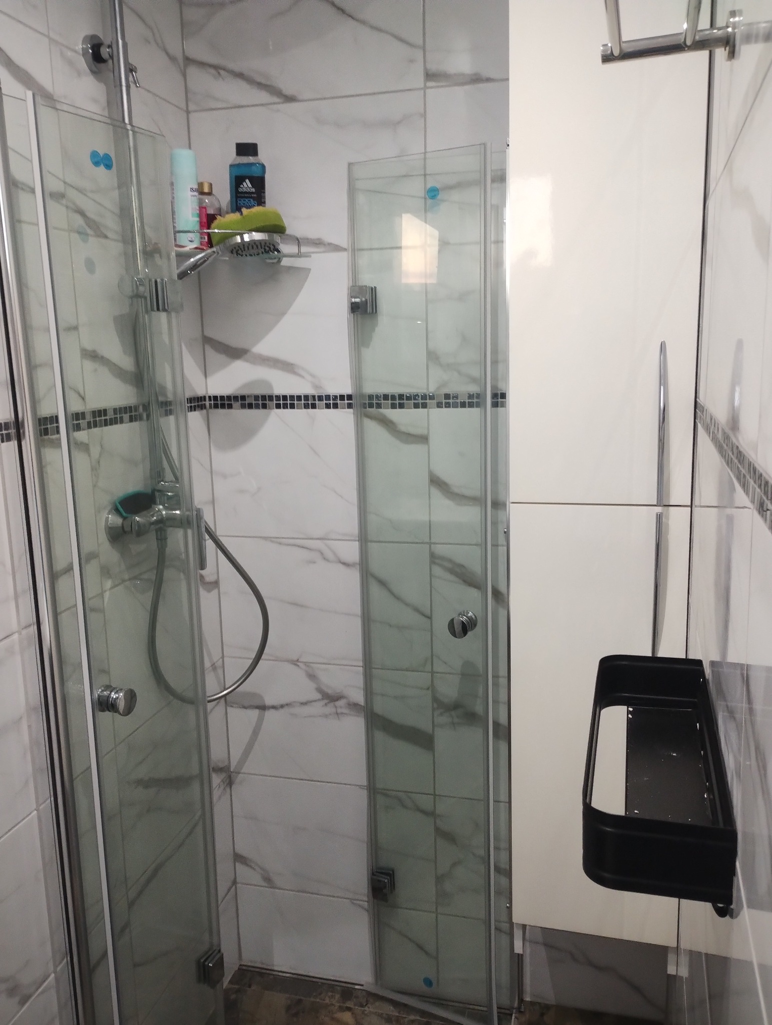 Nowoczesna kabina prysznicowa z szklanymi drzwiami, marmurowe płytki z dekoracyjnym paskiem mozaiki, kosmetyki na półce i czarna półka na akcesoria prysznicowe.