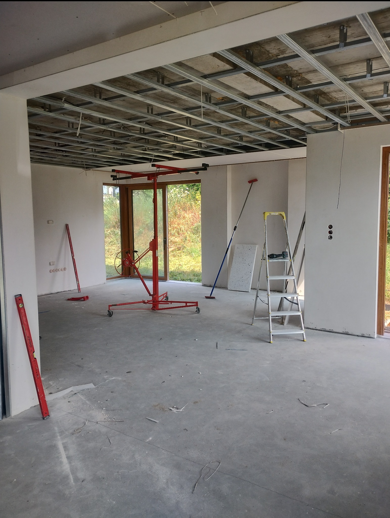 Wnętrze w trakcie montażu metalowego rusztu sufitowego podwieszanego, widoczny podnośnik do płyt, poziomice i drabina, surowy betonowy podkład.