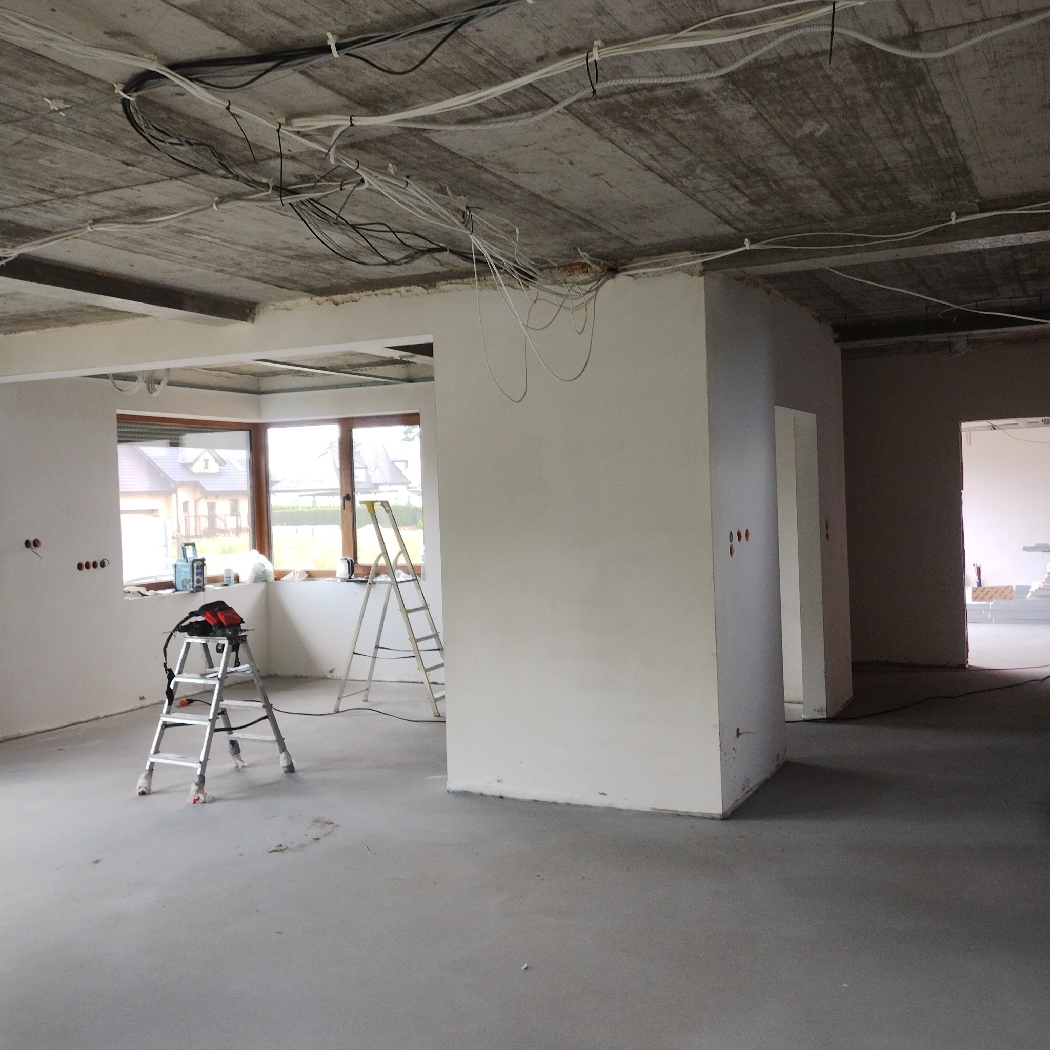 Surowy stan wnętrza w trakcie prac: widoczne kable pod sufitem, ściany przygotowane pod malowanie, dwie drabiny, otwory pod gniazdka elektryczne, posadzka betonowa.