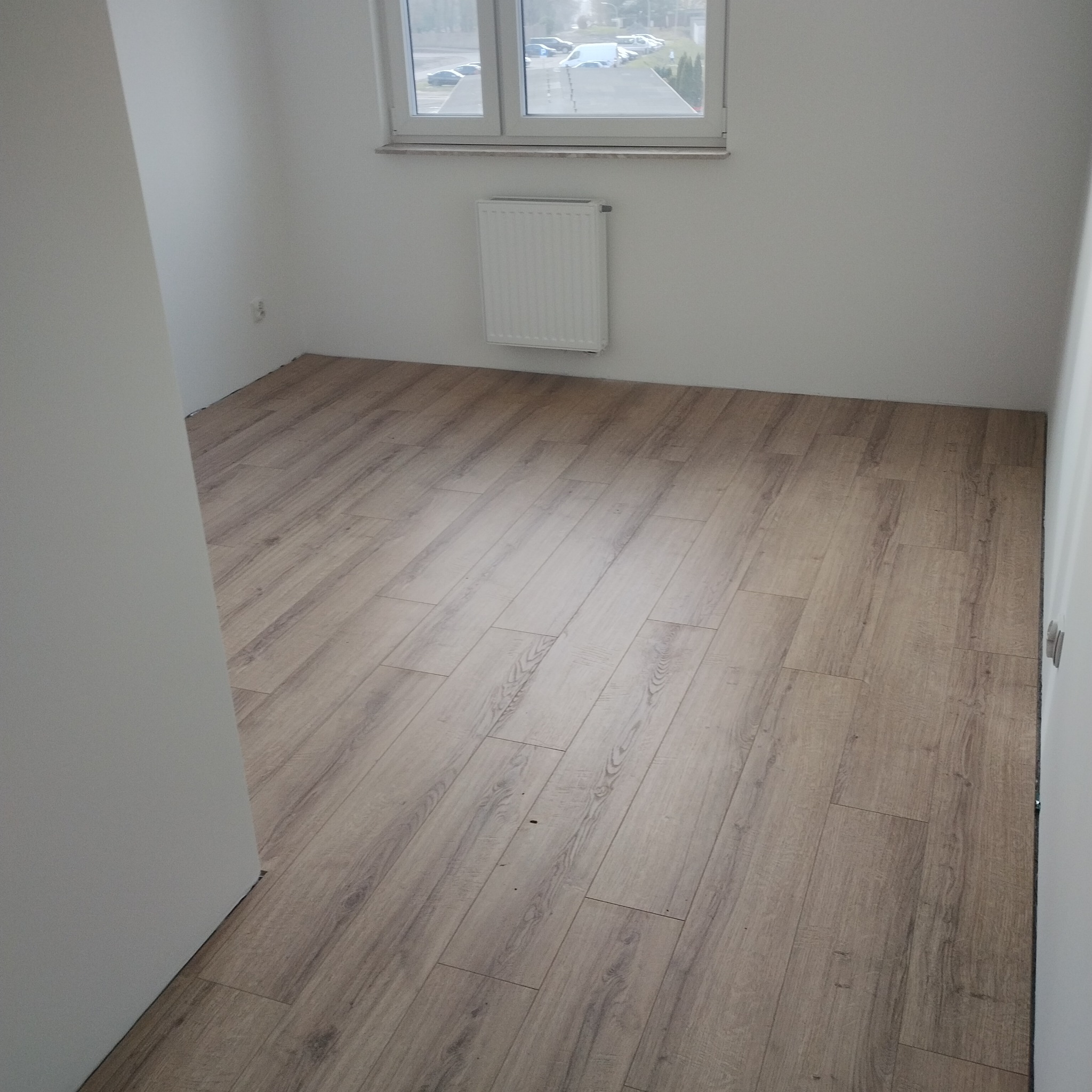 Jasne panele podłogowe ułożone w pustym pokoju z białymi ścianami i grzejnikiem pod oknem.