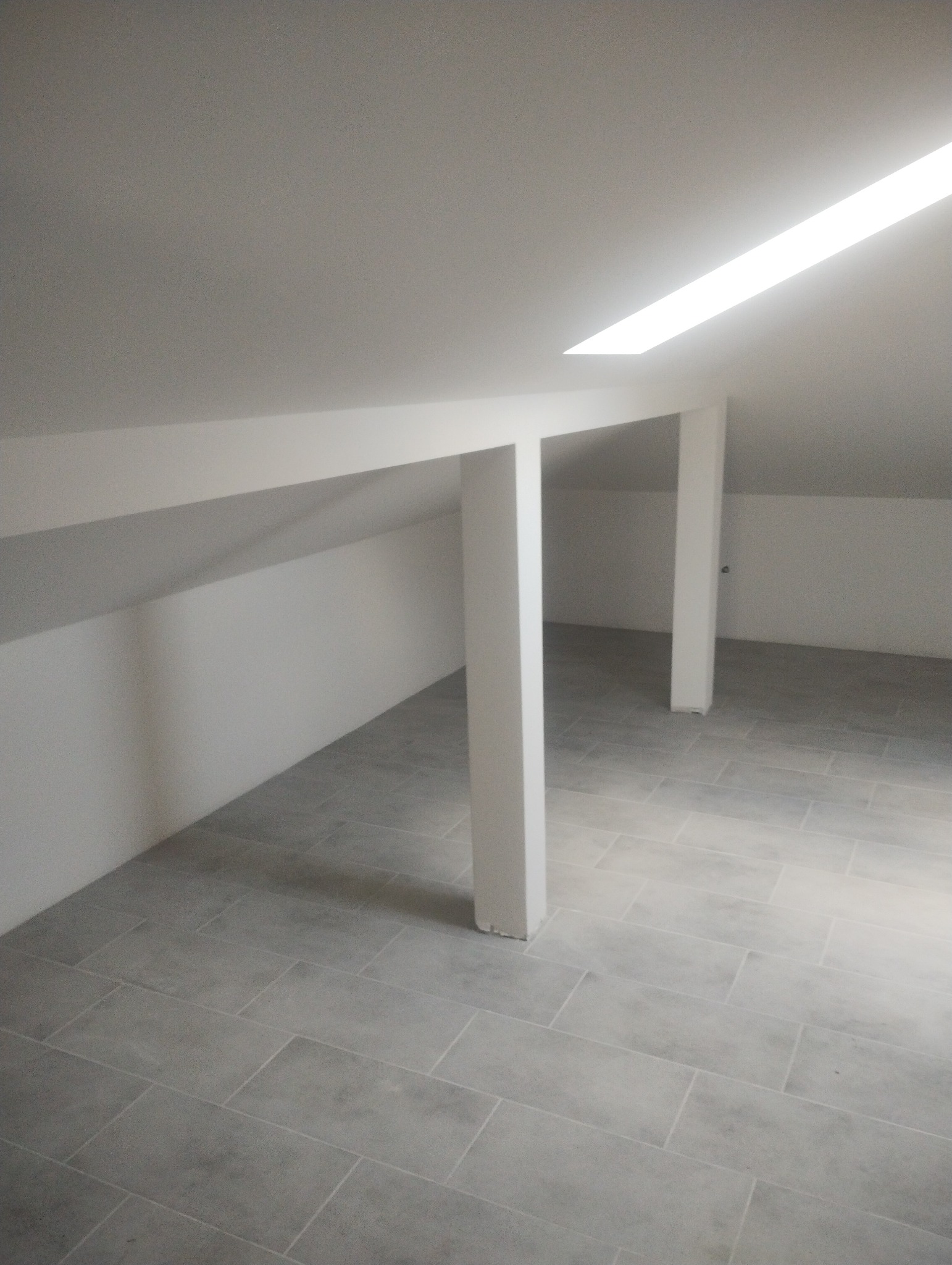 Poddasze po remoncie, widoczne białe ściany ze skosami, szare płytki podłogowe, białe filary podtrzymujące dach i wbudowane oświetlenie sufitowe.