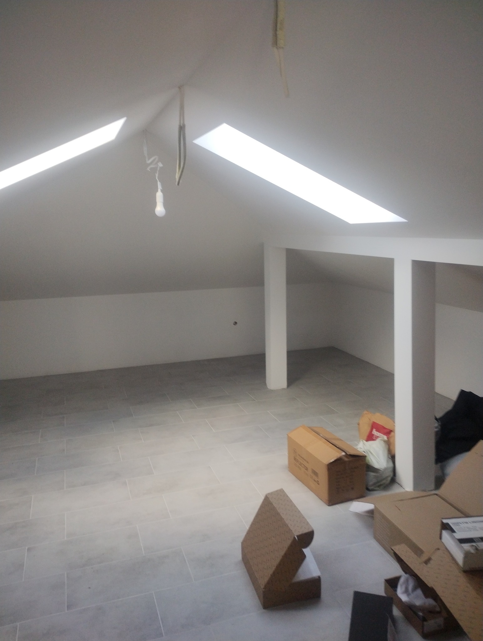 Poddasze z nową, jasnoszarą podłogą z płytek, widoczne kartony z materiałami budowlanymi, surowe ściany i okna dachowe.
