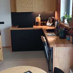 Artur Nowak - Nowoczesna kuchnia z drewnianymi frontami szafek górnych i czarnymi dolnymi, blat roboczy z jasnego drewna, widoczne akcesoria kuchenne i stół z ludowym obrusem.