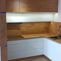 Artur Nowak - Nowoczesna kuchnia z białymi szafkami dolnymi, drewnopodobnymi szafkami górnymi i blatem, z wbudowanym piekarnikiem i mikrofalówką, oraz czarną armaturą. Widoczne okno z żaluzjami.