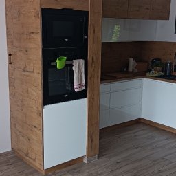 Artur Nowak - Nowoczesna zabudowa kuchenna z piekarnikiem i mikrofalówką Beko, fronty w kolorze drewna i białym, z drewnianą kolumną i podłogą z paneli imitujących drewno.