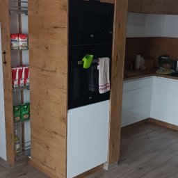 Artur Nowak - Nowoczesna kuchnia z drewnianymi frontami, zabudowana piekarnikiem i wysuwaną szafką cargo z zapasami, całość na drewnopodobnej podłodze.