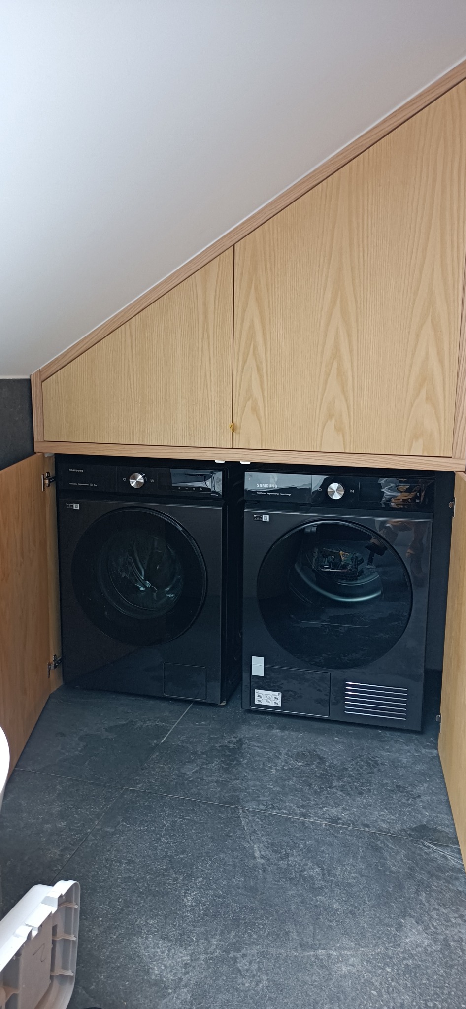 Dwie czarne pralki automatyczne Samsung umieszczone obok siebie pod zabudową z jasnego drewna w skosie, szara podłoga z płytek.