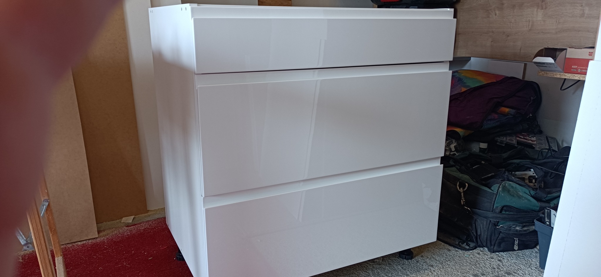 Biała komoda z trzema szufladami o wysokim połysku, widoczna w surowym pomieszczeniu z narzędziami i materiałami budowlanymi w tle.