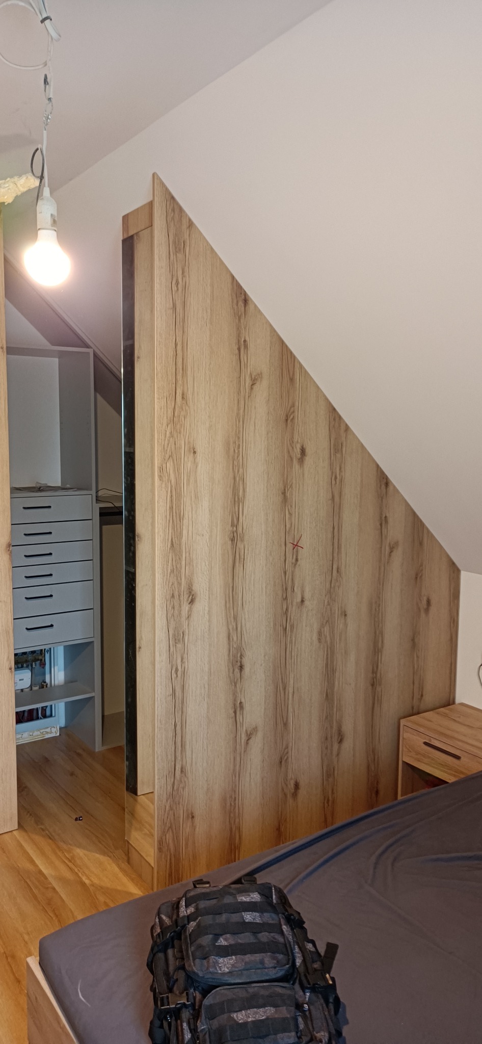 Zabudowa wnęki na poddaszu z drewnopodobną okleiną, widoczny fragment szafy z szufladami wewnątrz i plecak na łóżku.