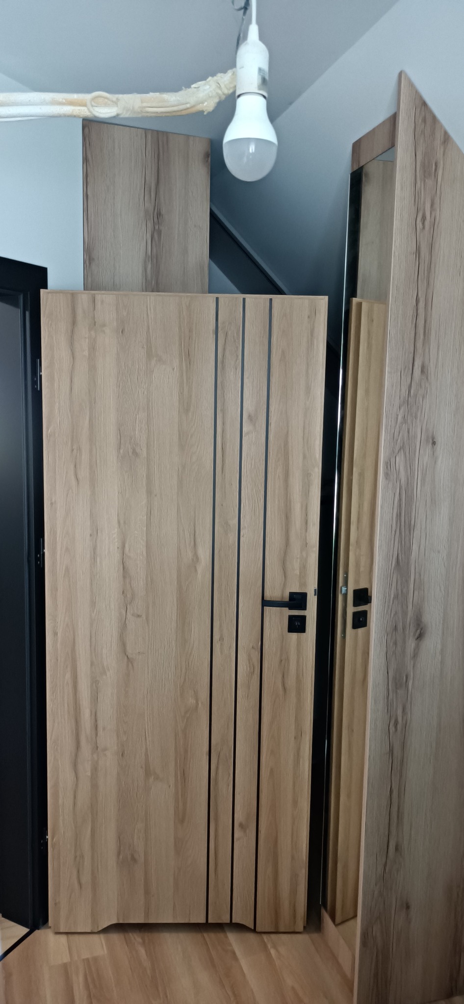 Fragment szafy z drewnopodobnymi frontami i czarnymi, pionowymi wstawkami, widoczna czarna klamka i ościeżnica drzwi po lewej stronie, w tle sufit z lampą.