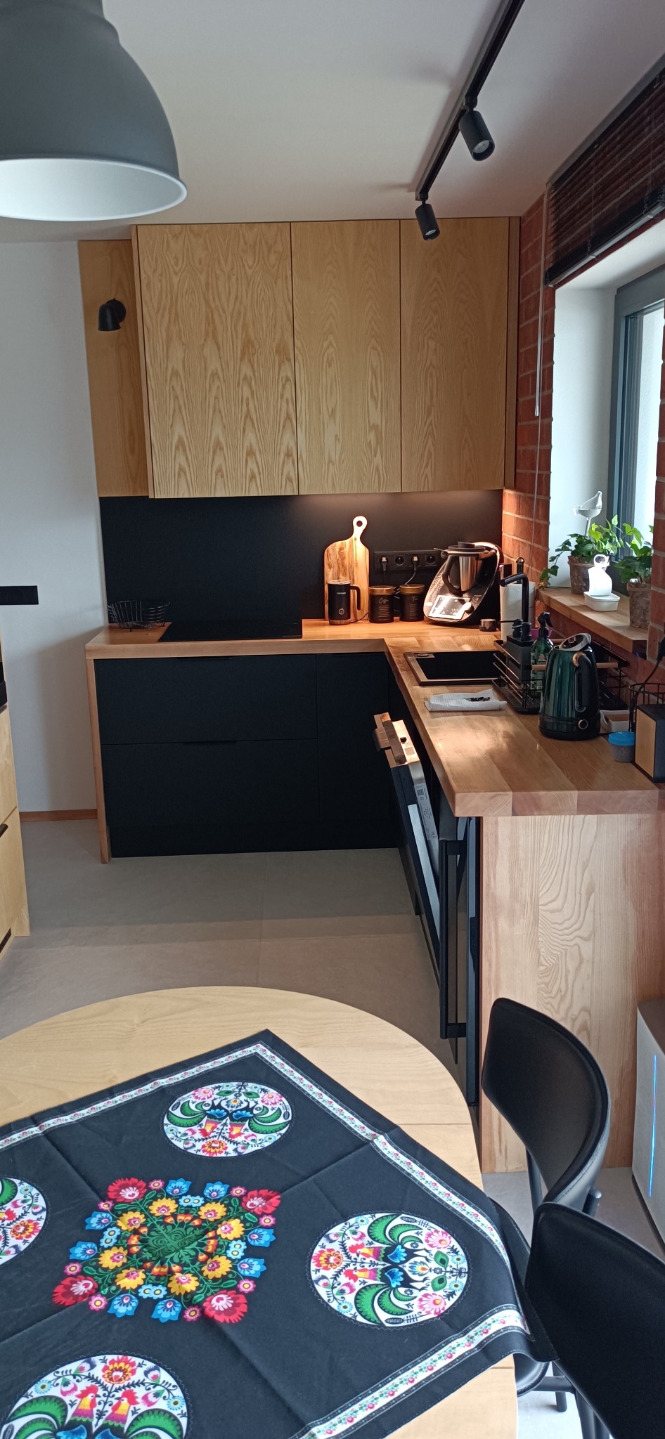 Nowoczesna kuchnia z drewnianymi frontami szafek górnych i czarnymi dolnymi, blat roboczy z jasnego drewna, widoczne akcesoria kuchenne i stół z ludowym obrusem.