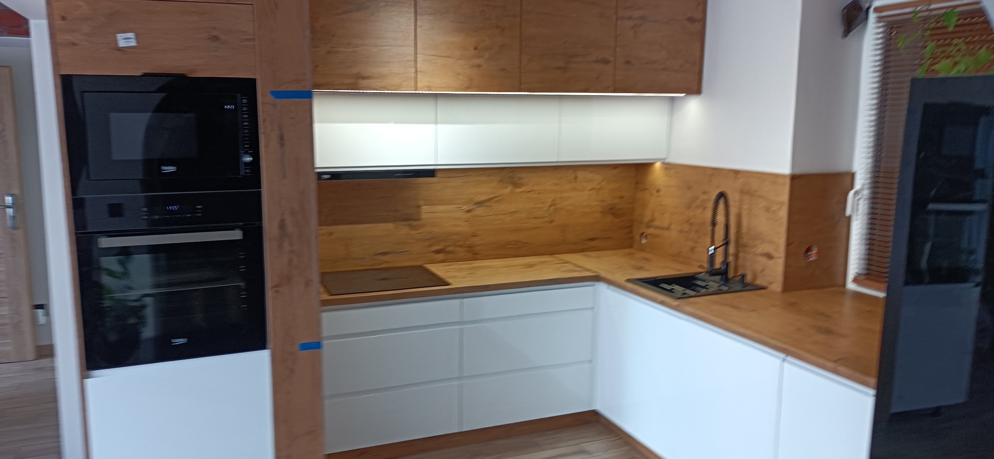 Nowoczesna kuchnia z białymi szafkami dolnymi, drewnopodobnymi szafkami górnymi i blatem, z wbudowanym piekarnikiem i mikrofalówką, oraz czarną armaturą. Widoczne okno z żaluzjami.