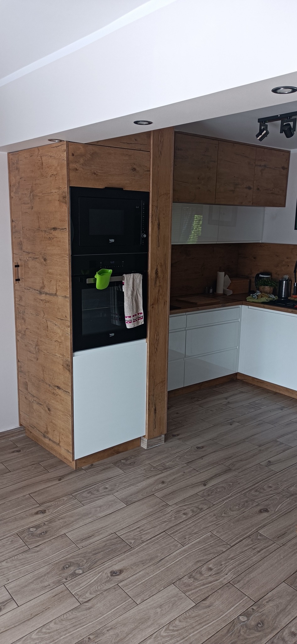 Nowoczesna zabudowa kuchenna z piekarnikiem i mikrofalówką Beko, fronty w kolorze drewna i białym, z drewnianą kolumną i podłogą z paneli imitujących drewno.
