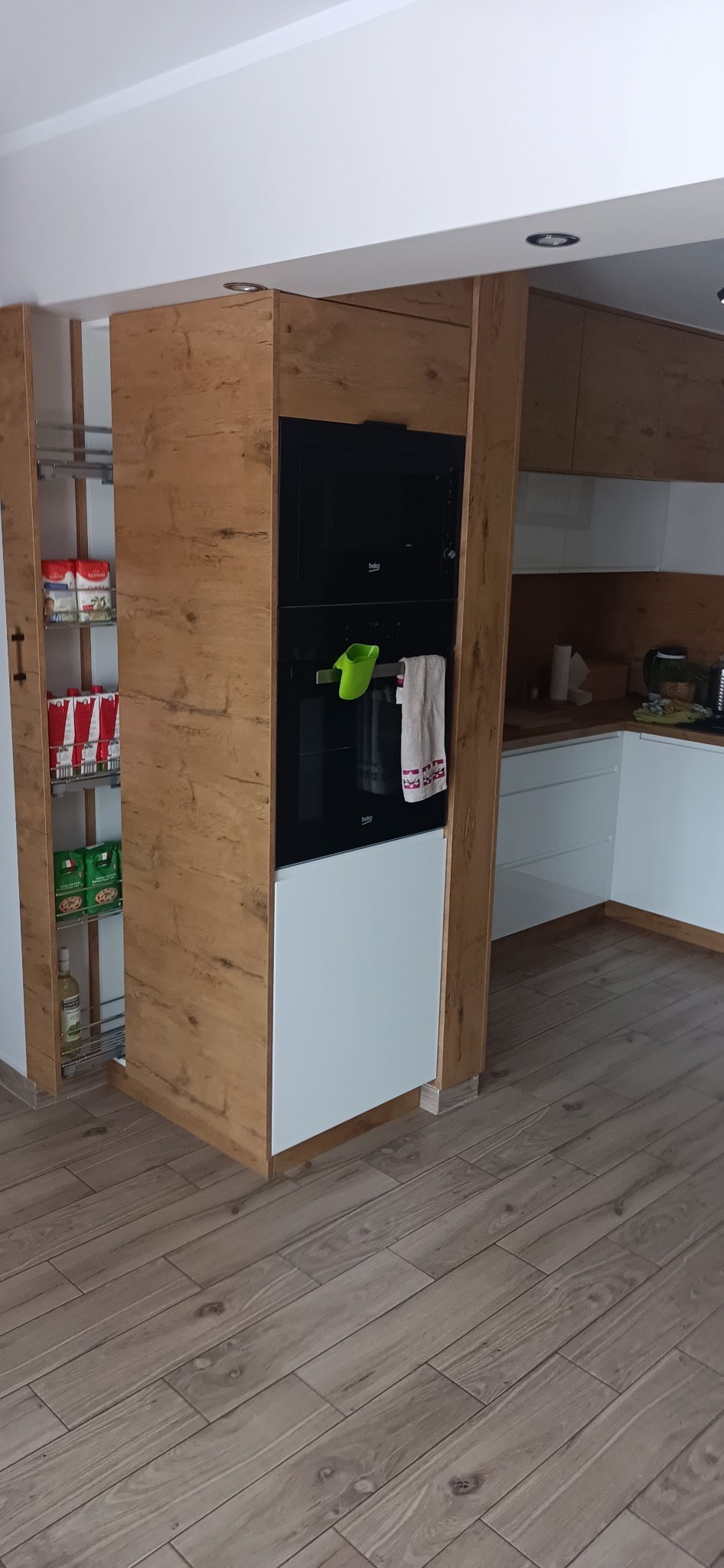 Nowoczesna kuchnia z drewnianymi frontami, zabudowana piekarnikiem i wysuwaną szafką cargo z zapasami, całość na drewnopodobnej podłodze.