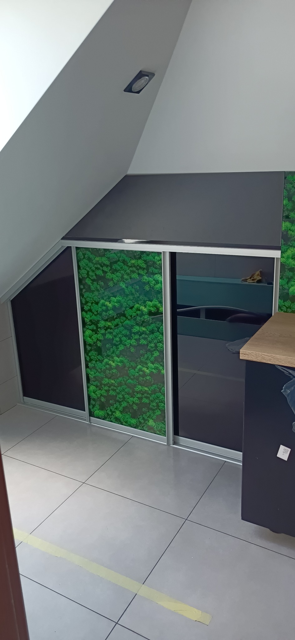 Szafa wnękowa z przesuwnymi drzwiami, z jednym skrzydłem w kolorze czarnym i drugim z fototapetą przedstawiającą las, zamontowana na poddaszu.