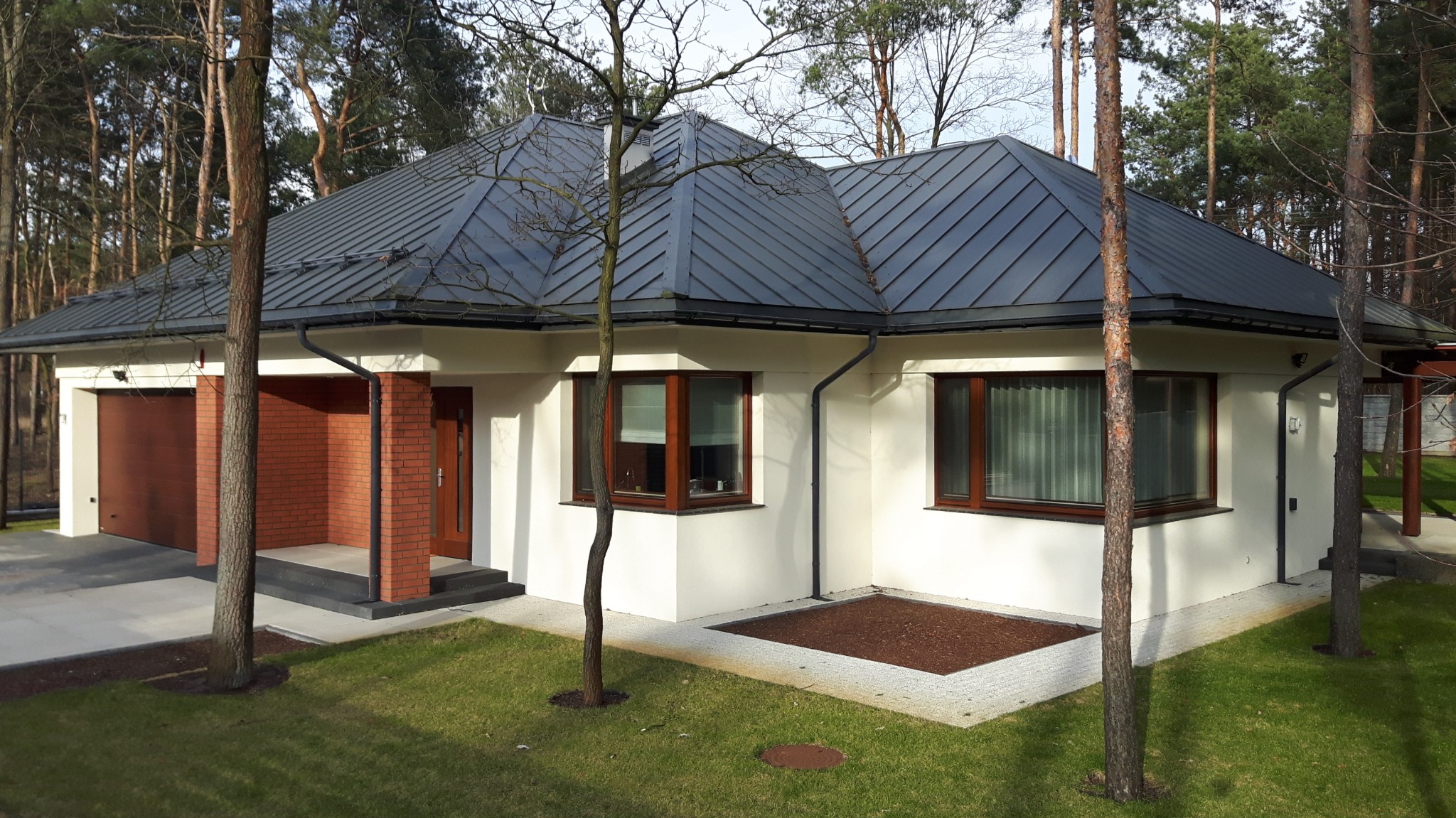 Nowoczesny dom murowany z ciemnoszarą blachodachówką na tle lasu, z garażem po lewej stronie i wejściem z ceglaną wnęką pośrodku.