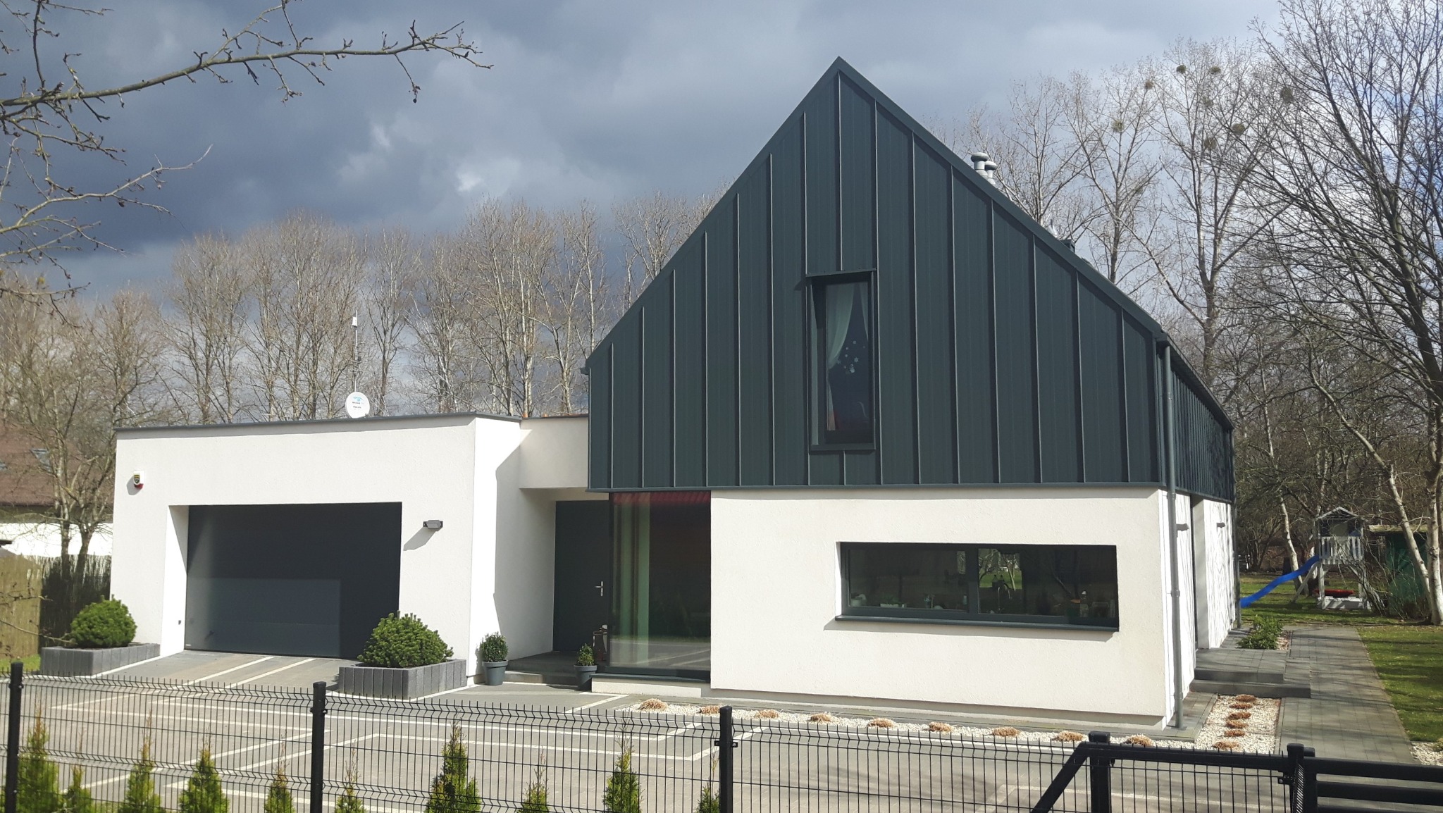 Nowoczesny dom murowany z grafitową blachą na dachu i elewacją w kolorze złamanej bieli, widoczny zza ogrodzenia z metalowych paneli, na tle pochmurnego nieba i drzew bez liści.