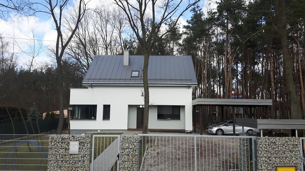 Nowoczesny, parterowy dom z szarą dachówką i podwójnym carportem w Legionowie, otoczony drzewami, widziany zza ogrodzenia z gabionów.