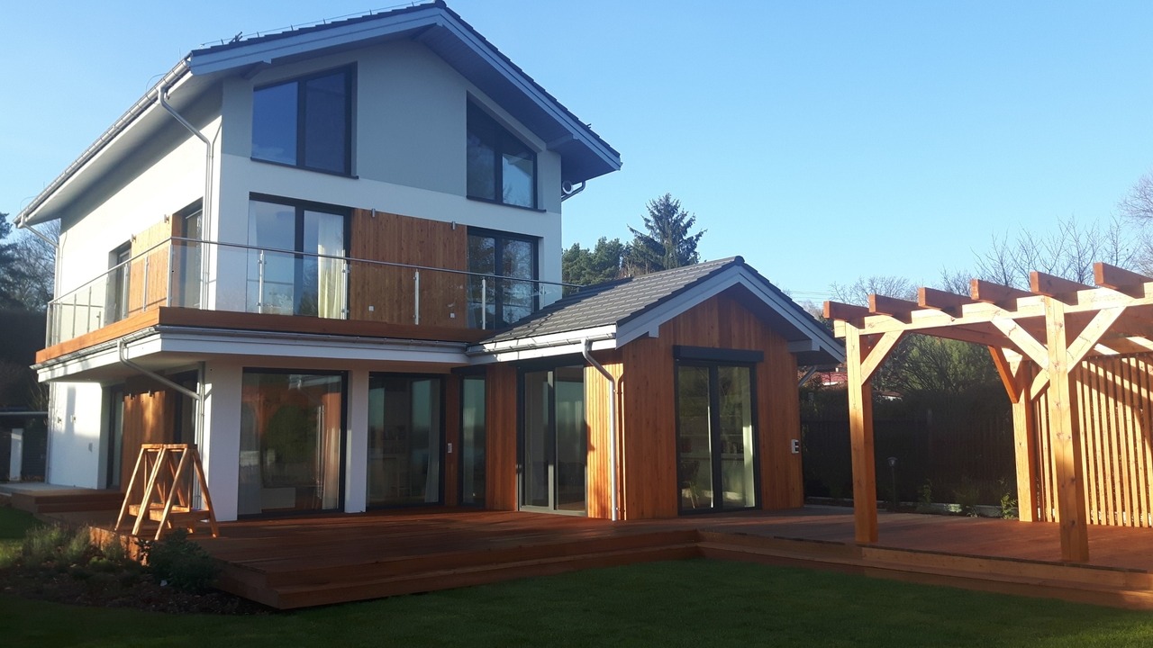 Dwukondygnacyjny dom z drewnianą elewacją, balkonem ze szklaną balustradą i tarasem z drewnianą pergolą w słoneczny dzień.