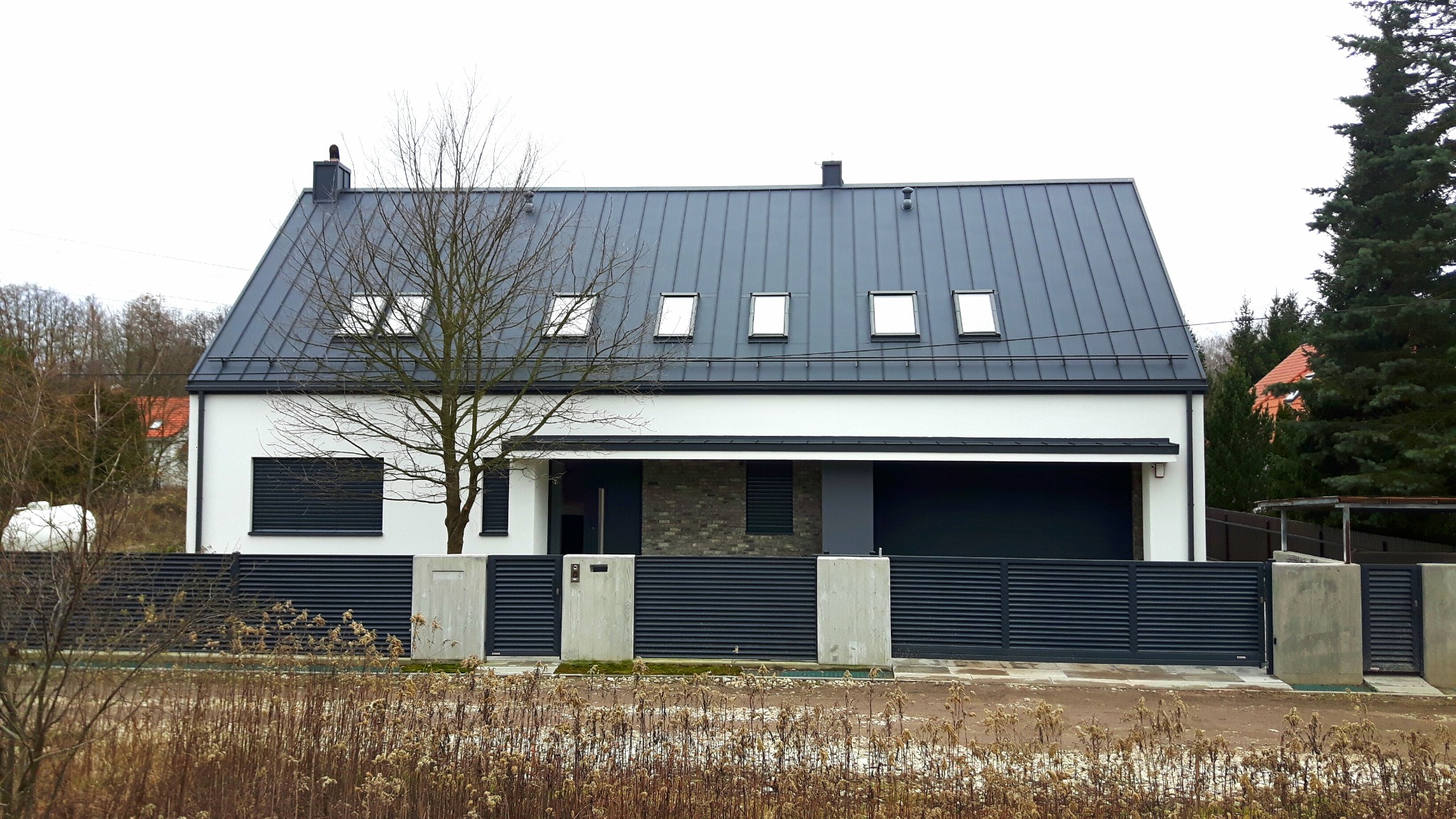 Nowoczesny dom jednorodzinny z szarą blachodachówką, białymi ścianami i grafitowym ogrodzeniem lamelowym, widok z zewnątrz w pochmurny dzień.