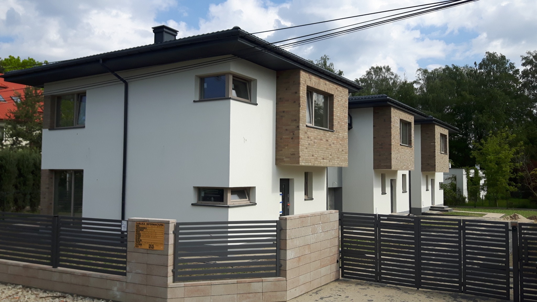 Dwa nowoczesne, bliźniacze domy z białymi ścianami i ceglanymi elementami, ogrodzone metalowym płotem z ceglanymi słupkami, z tablicą informacyjną po lewej stronie.