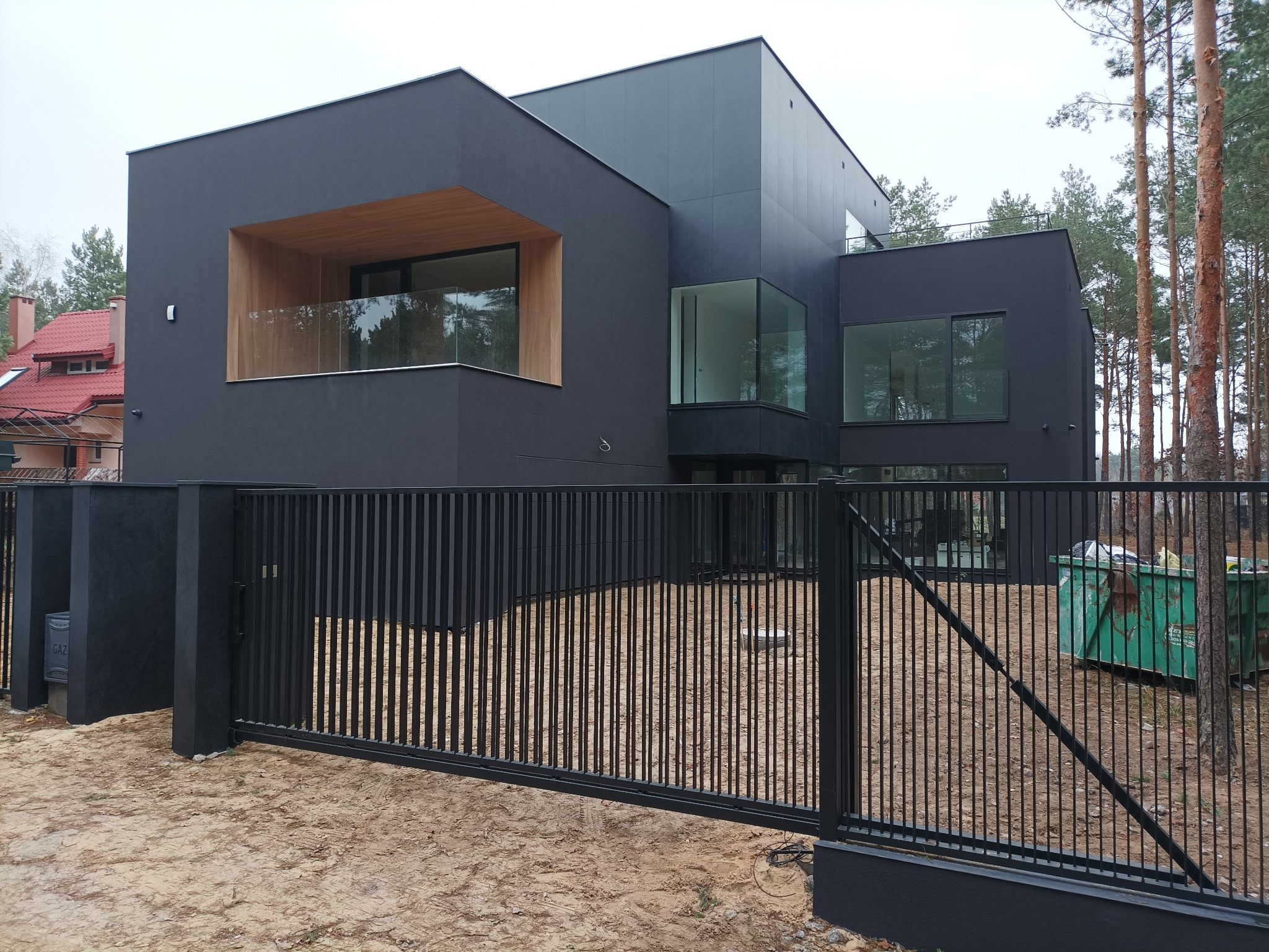 Nowoczesny, dwukondygnacyjny dom o kubistycznej architekturze, pomalowany na ciemny kolor z drewnianymi akcentami na balkonie i dużymi oknami, otoczony czarnym, metalowym ogrodzeniem i zielonym...