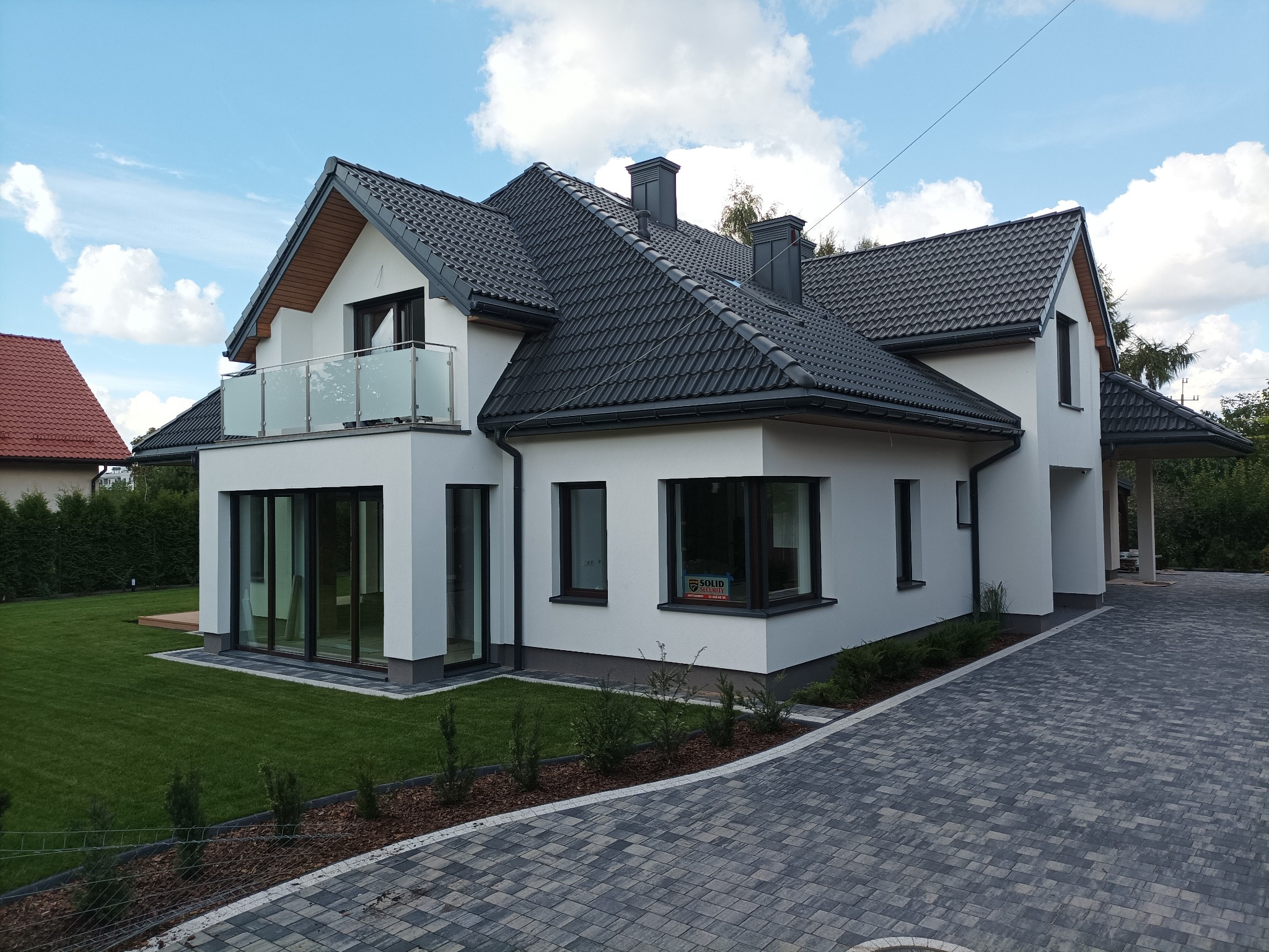 Nowoczesny dom z białymi ścianami, ciemną dachówką i szklaną balustradą balkonu, widok z poziomu brukowanego podjazdu.