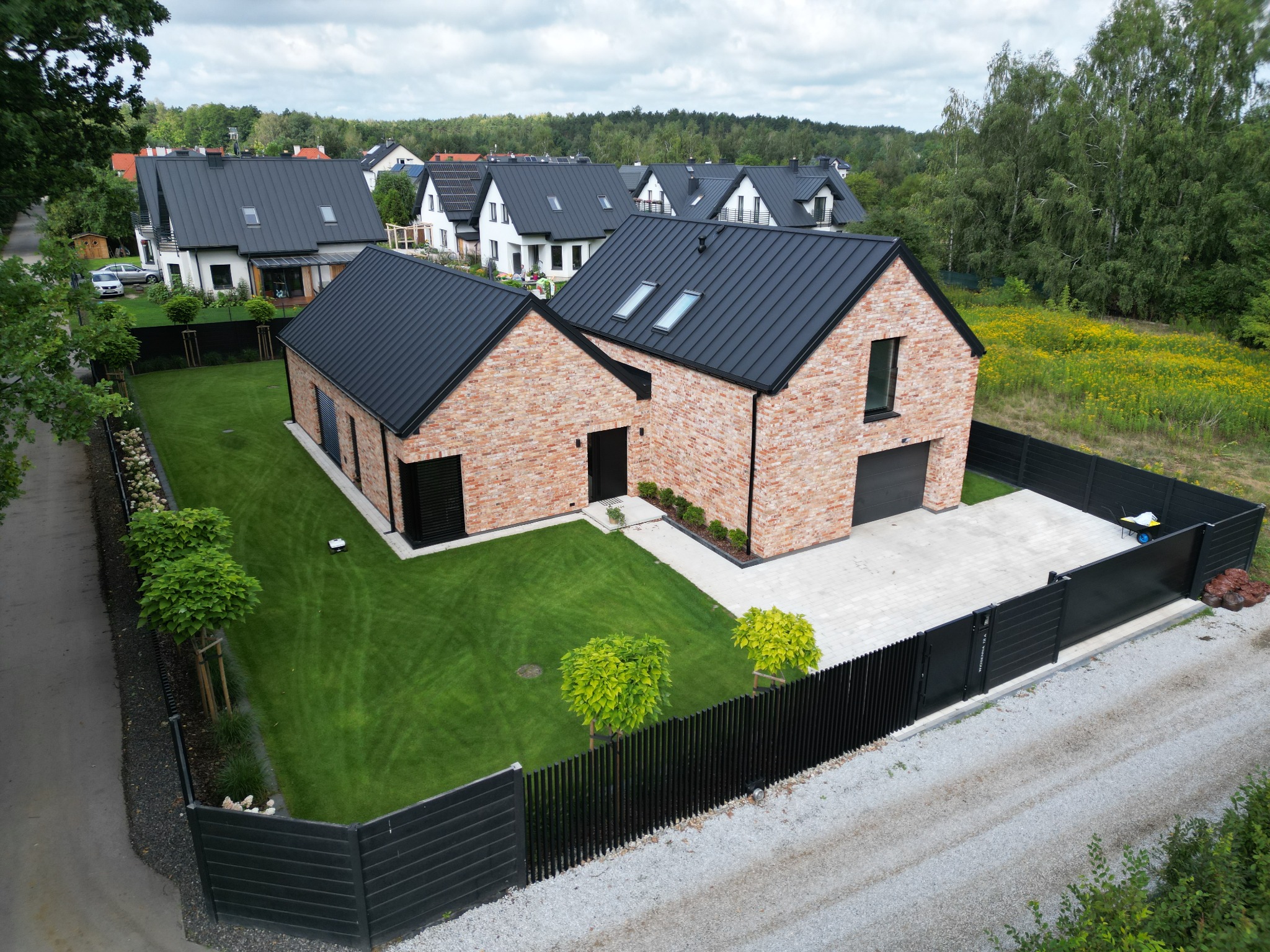 Nowoczesny dom jednorodzinny z cegły klinkierowej z czarnym dachem na tle innych domów i zieleni, widok z góry.