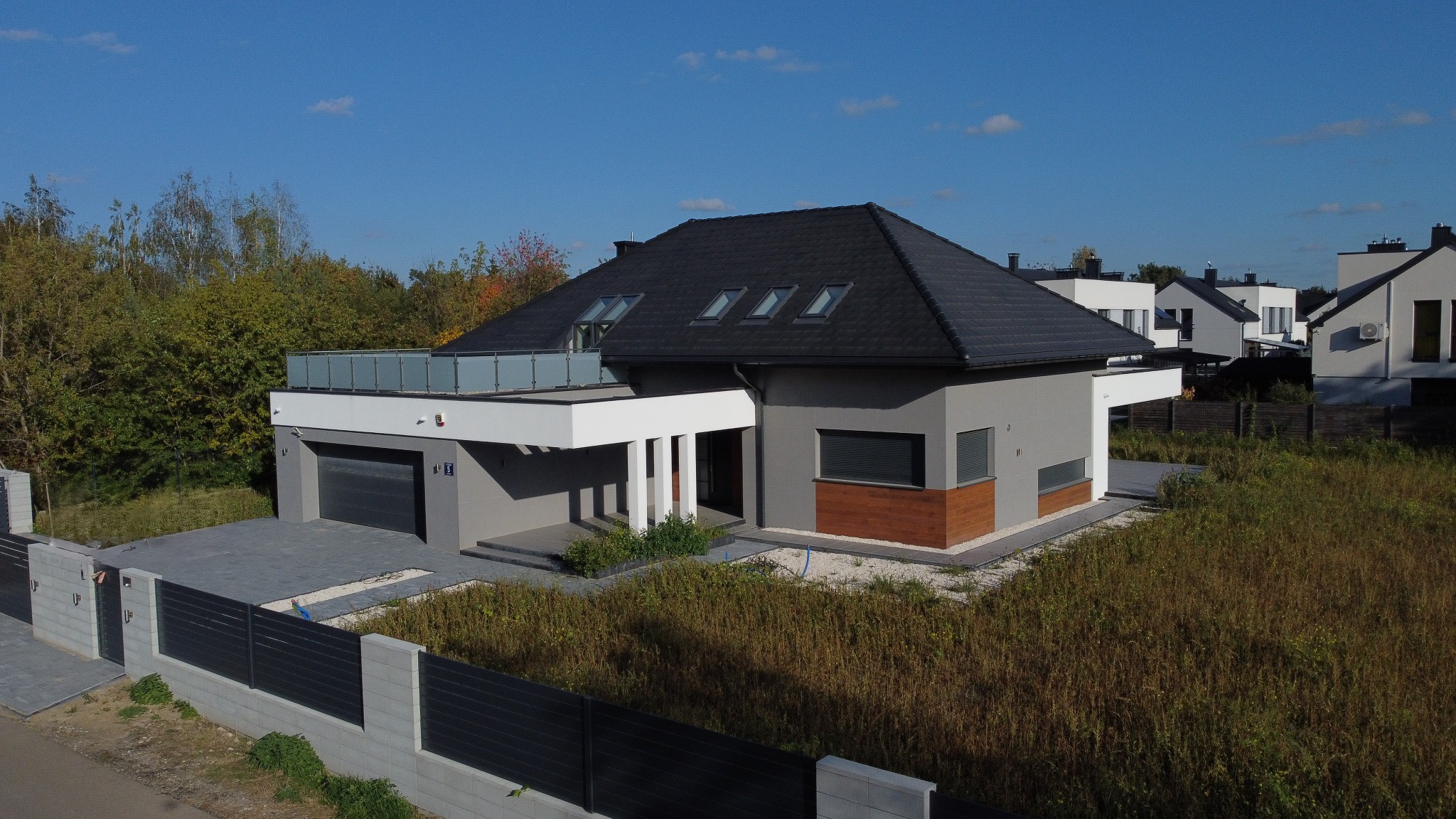 Nowoczesny dom jednorodzinny z płaskim dachem i balkonem z balustradą ze szkła, widok z góry, szara elewacja z drewnianymi akcentami, czarna dachówka, trzy okna dachowe, brama garażowa segmentowa...