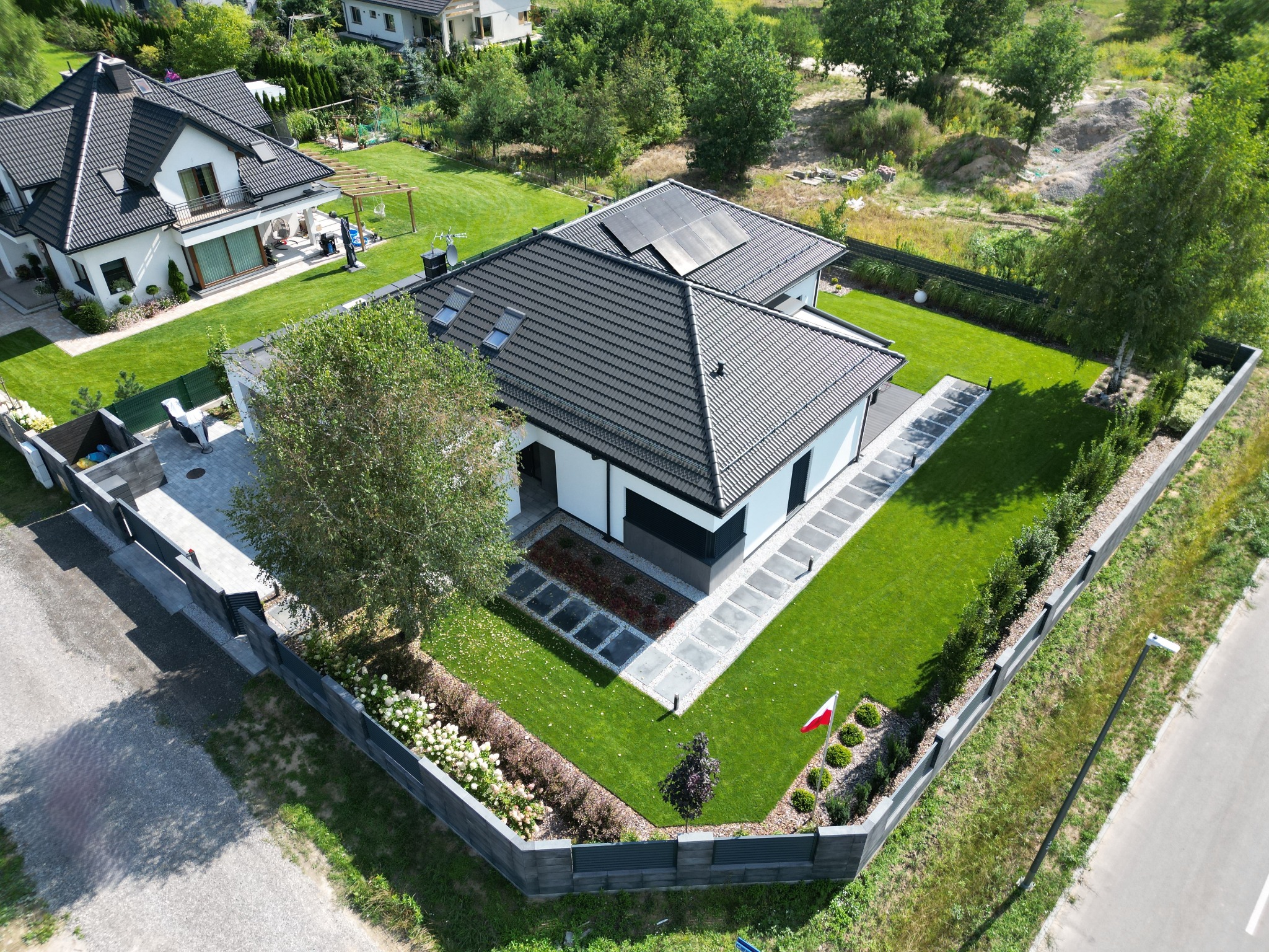 Nowoczesny dom z ciemną dachówką i panelami słonecznymi, widok z lotu ptaka, starannie zagospodarowany ogród z kostką brukową i elementami dekoracyjnymi, ogrodzenie z polską flagą w rogu działki.