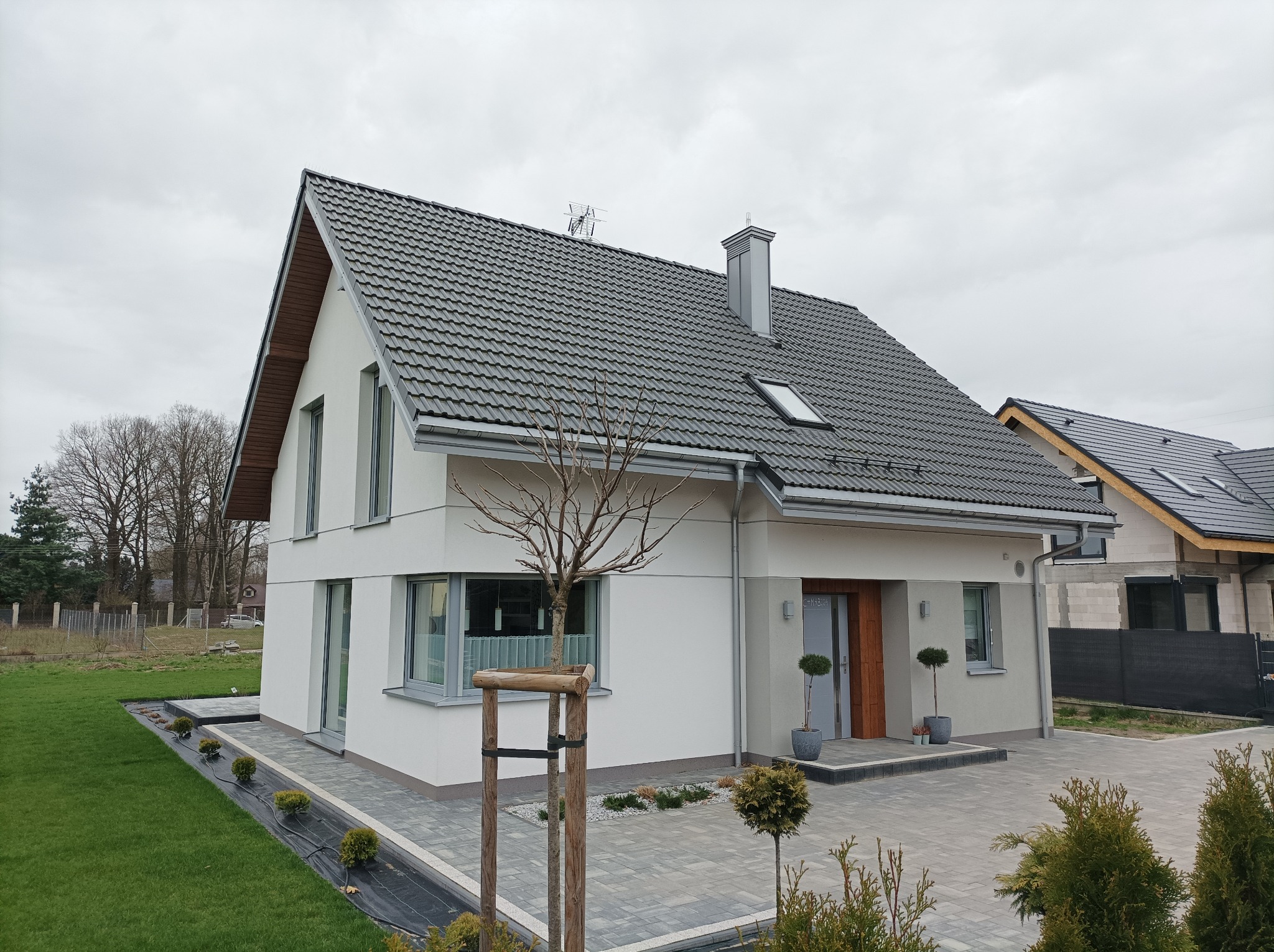 Nowoczesny dom jednorodzinny z szarą dachówką, jasną elewacją i starannie zagospodarowanym terenem wokół, w tym wybrukowany podjazd i świeżo posadzona zieleń.