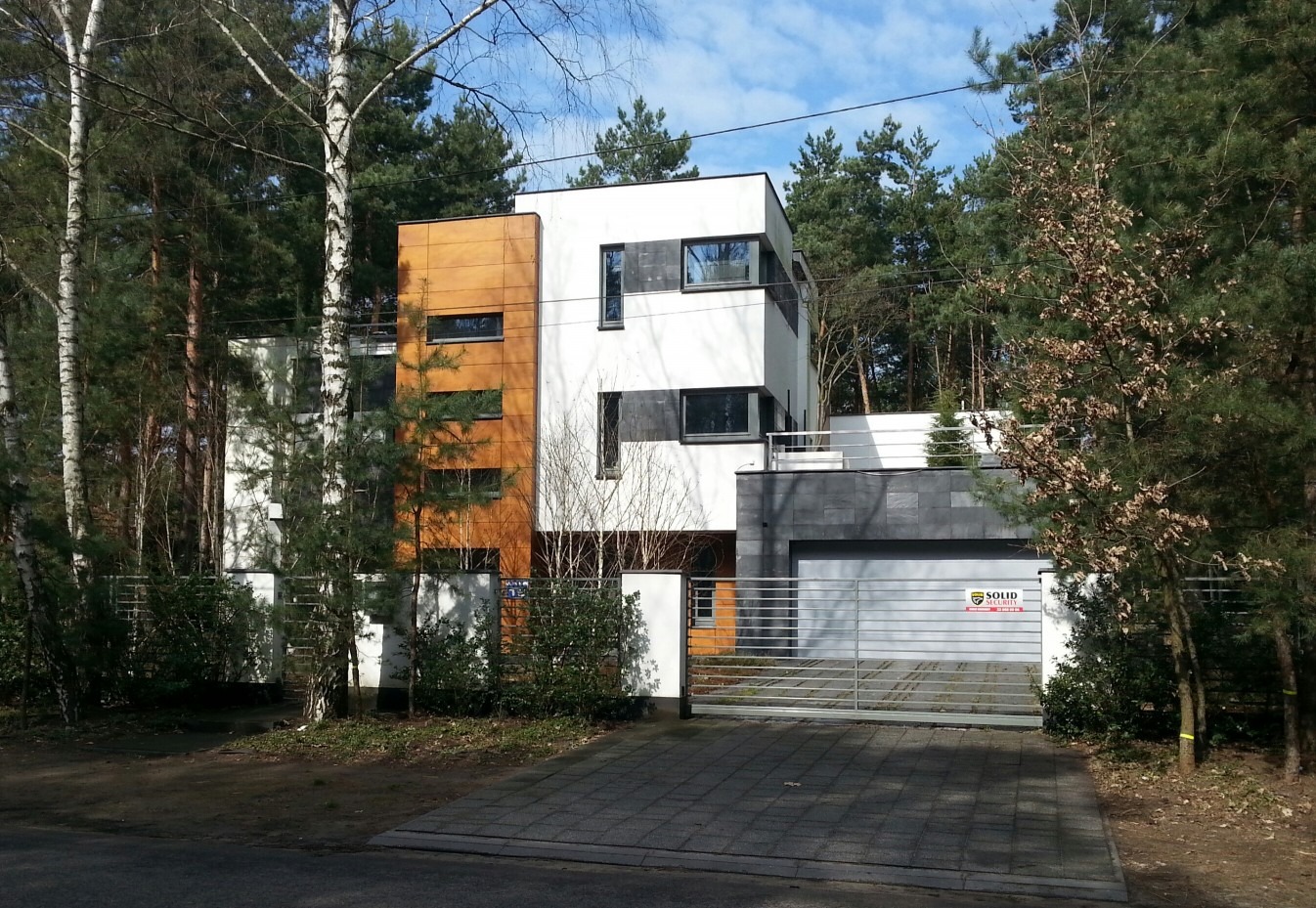 Nowoczesny dom murowany z elementami drewna i szarej elewacji, widoczny z zewnątrz przez metalową bramę wjazdową i ogrodzenie, otoczony drzewami.