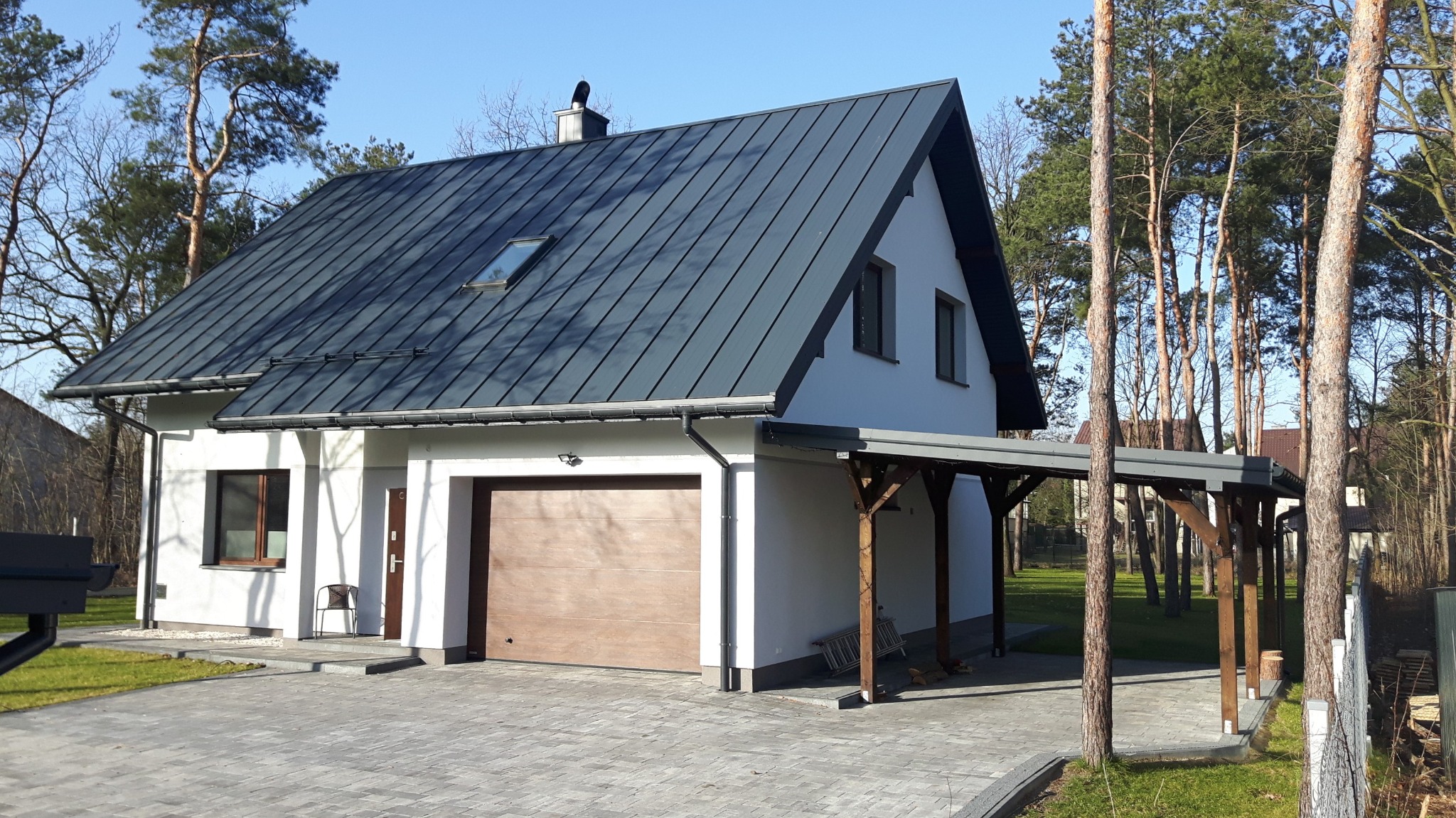 Nowoczesny dom jednorodzinny z ciemnoszarą blachodachówką, drewnianą wiatą garażową i podjazdem z kostki brukowej, otoczony drzewami.