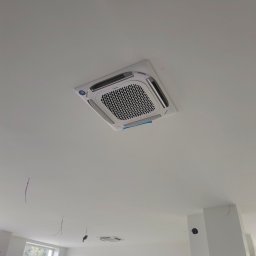InstalBlu - Kasetonowa jednostka klimatyzacyjna Midea zamontowana w suficie podwieszanym, widoczne niedokończone prace wykończeniowe i wystające przewody.