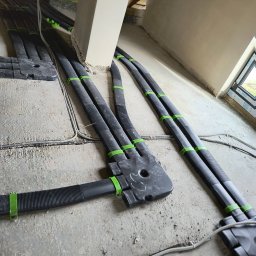 InstalBlu - Szczegółowe ujęcie instalacji przewodów wentylacyjnych z czarnych rur karbowanych, mocowanych zielonymi uchwytami, prowadzących wzdłuż surowej ściany poddasza w trakcie montażu.