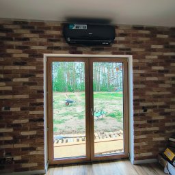 InstalBlu - Czarny klimatyzator ścienny zamontowany nad drewnianymi drzwiami balkonowymi w pokoju z ceglaną ścianą, widok na zewnątrz na trawnik z taczką.