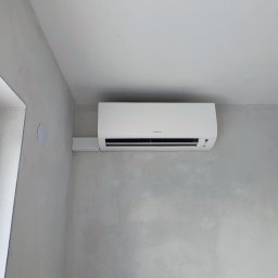 InstalBlu - Biała jednostka klimatyzacyjna Daikin zamontowana na surowej, szarej ścianie w nowym budynku, widoczny fragment okna po lewej stronie.