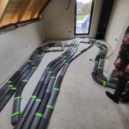 InstalBlu - Rozprowadzenie czarnych, elastycznych rur wentylacyjnych z zielonymi obejmami na poddaszu w trakcie budowy, widok z góry, fragment sylwetki osoby w kwiecistej bluzce.