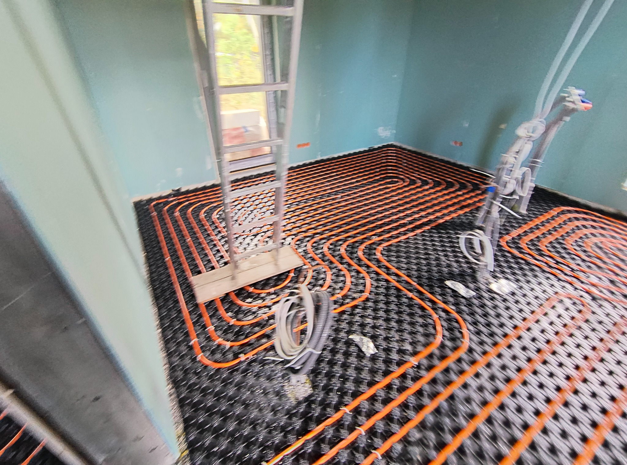 Pomieszczenie w trakcie instalacji ogrzewania podłogowego z pomarańczowymi rurkami na czarnej macie, widoczna drabina aluminiowa i przewody przyłączeniowe.