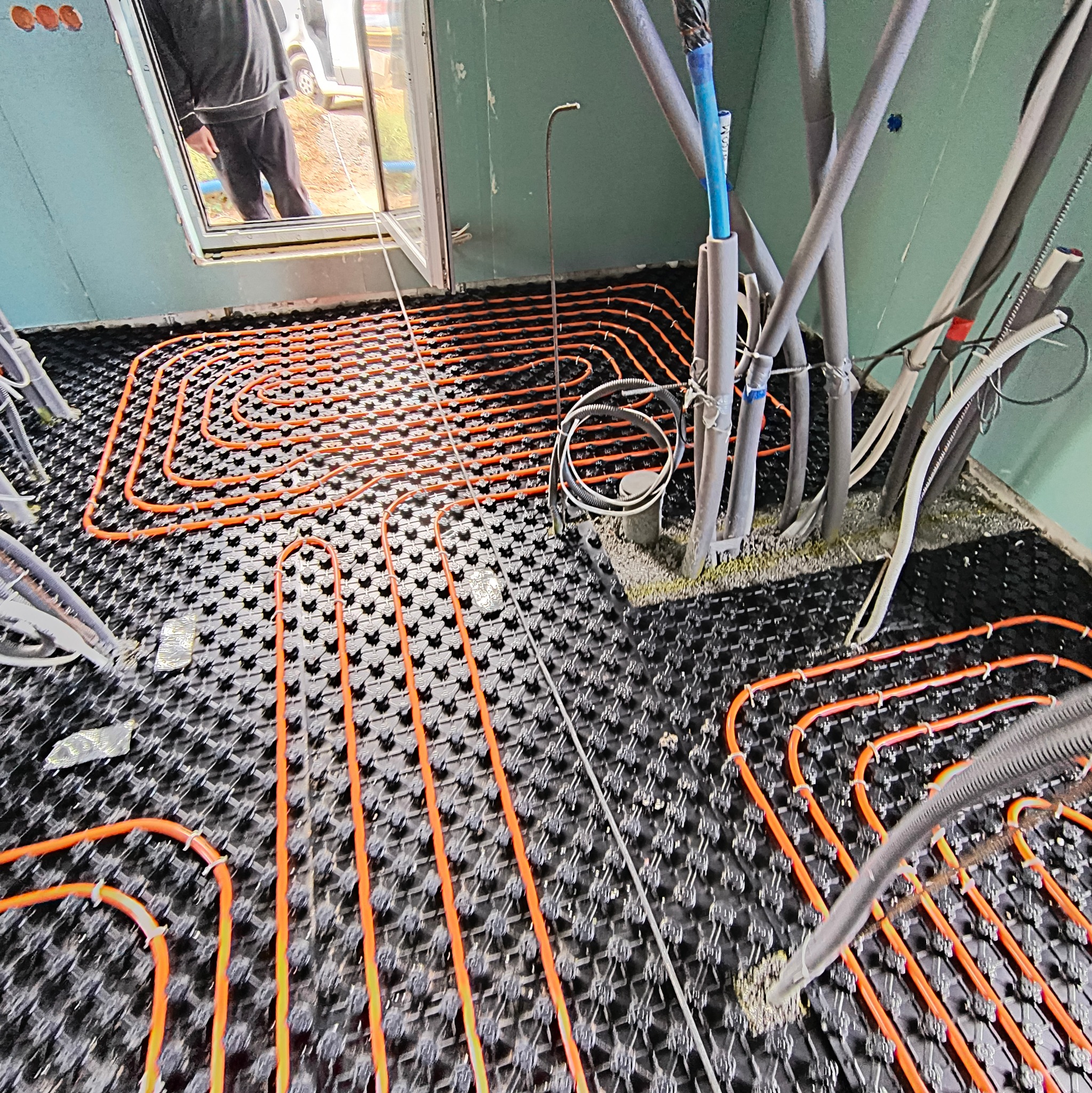 Pomarańczowe rury ogrzewania podłogowego ułożone na czarnej macie montażowej w surowym wnętrzu budynku, widoczne liczne kable i przewody oraz fragment okna z widokiem na zewnątrz.
