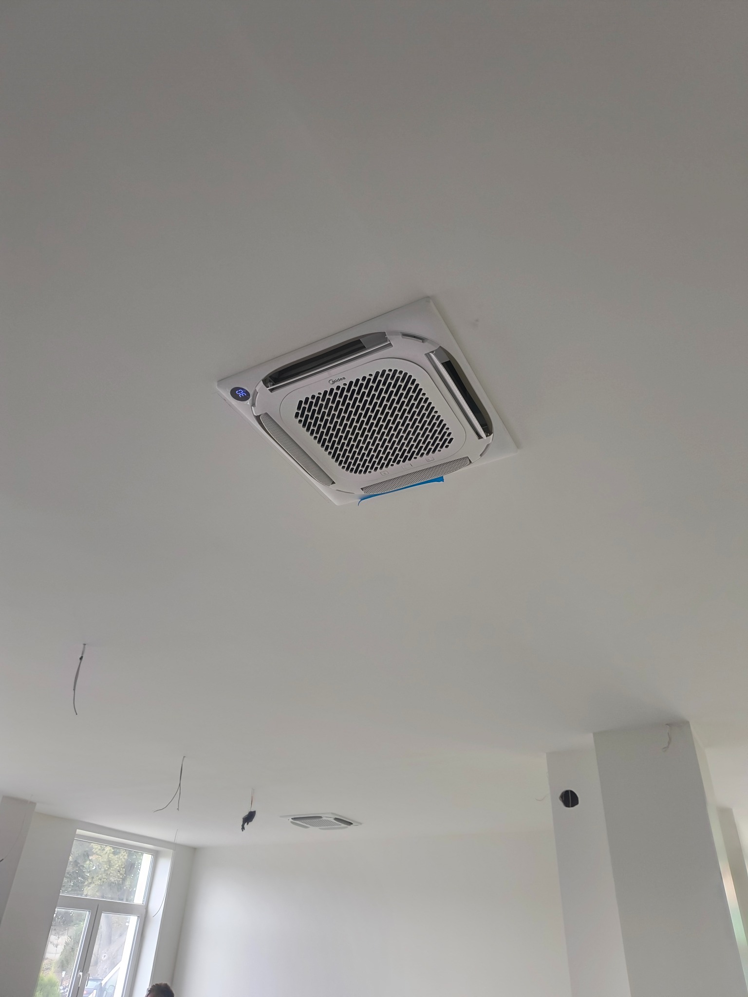 Kasetonowa jednostka klimatyzacyjna Midea zamontowana w suficie podwieszanym, widoczne niedokończone prace wykończeniowe i wystające przewody.