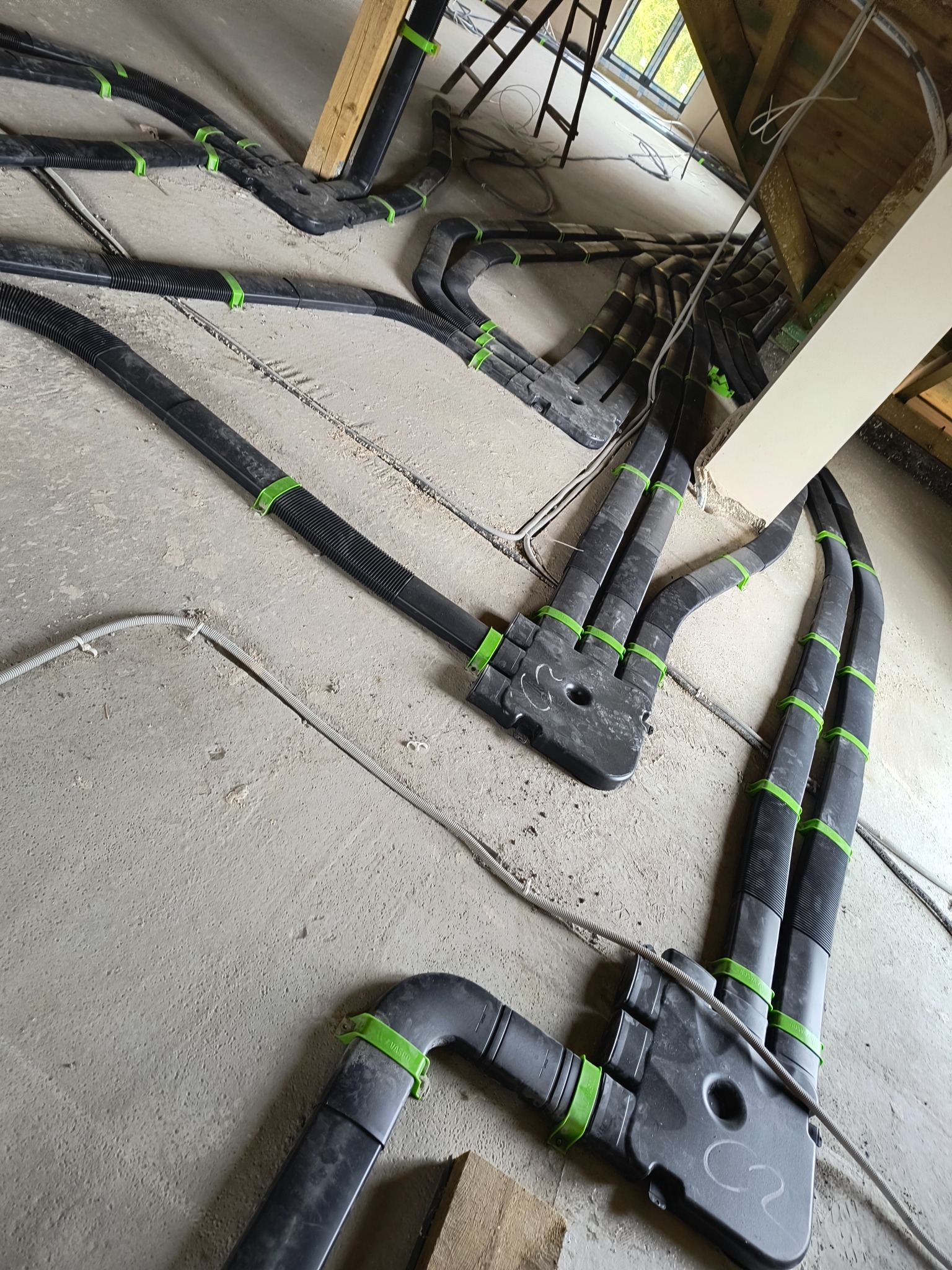 Rozprowadzenie czarnych, karbowanych rur wentylacyjnych z zielonymi opaskami mocującymi na poddaszu w Szubinie, widoczne elementy konstrukcji dachu i okno dachowe.