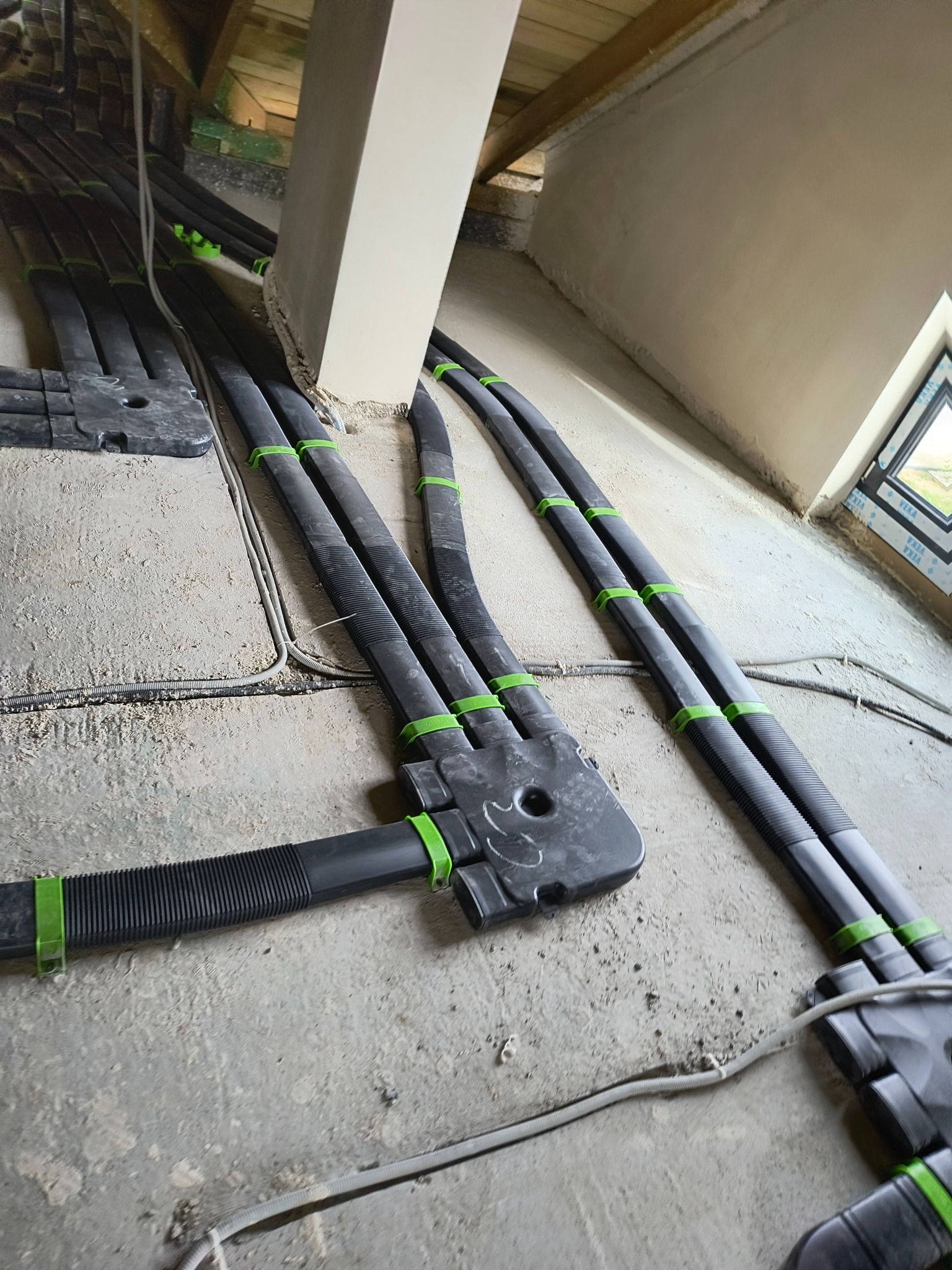 Szczegółowe ujęcie instalacji przewodów wentylacyjnych z czarnych rur karbowanych, mocowanych zielonymi uchwytami, prowadzących wzdłuż surowej ściany poddasza w trakcie montażu.